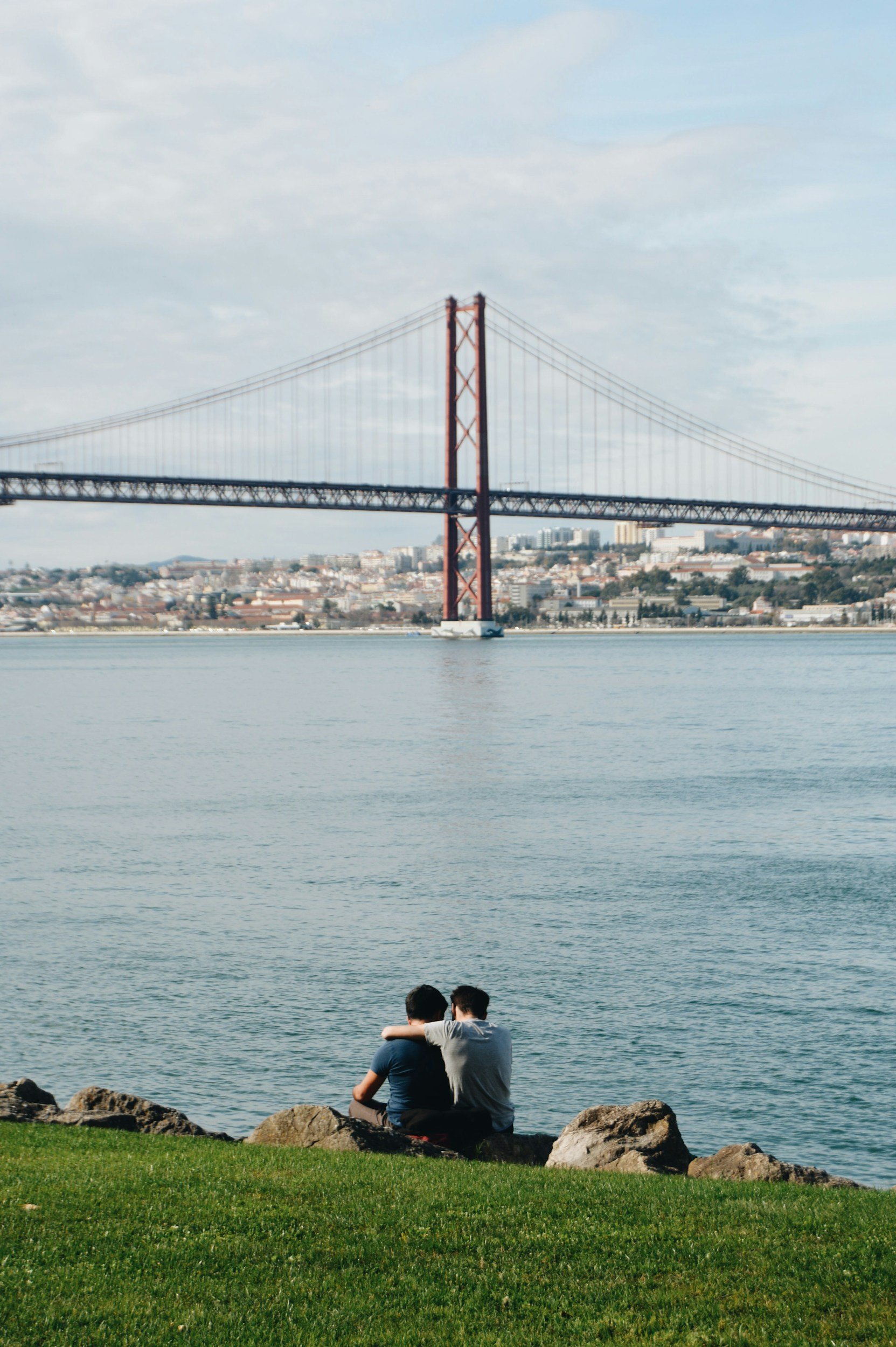 Two young men sit on rocks by the water, embracing while facing a large suspension bridge in the distance.
