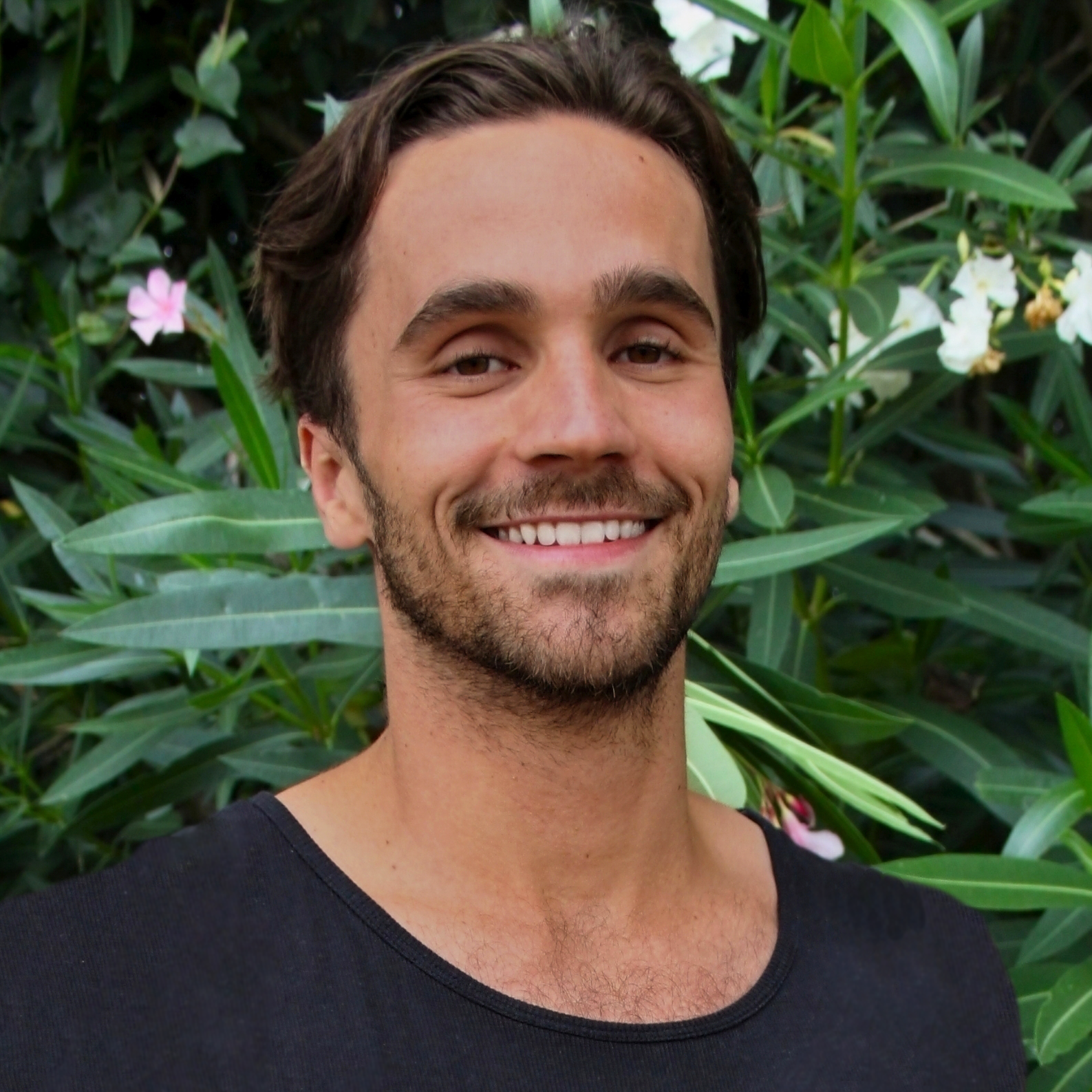 A smiling man with dark hair and a beard, wearing a black shirt, standing in front of green foliage with pink and white flowers.