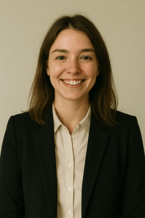 A young woman with shoulder-length brown hair, smiling, dressed in a black blazer and white shirt, standing against a beige wall.