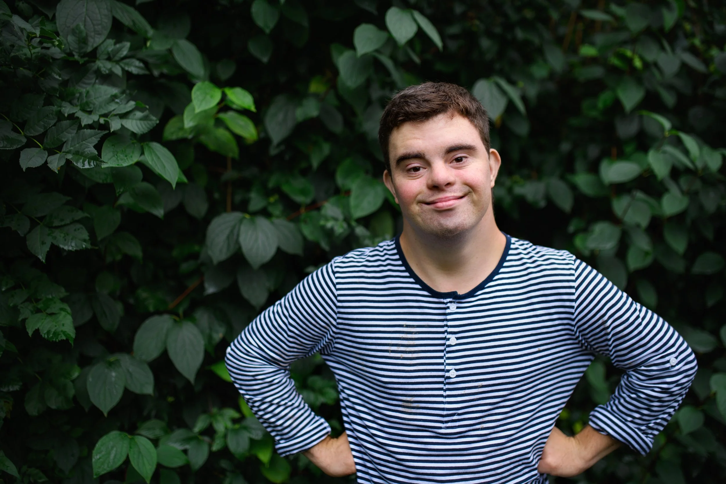 A young man with short brown hair, wearing a black and white striped shirt, stands with his hands on his hips in front of a dense backdrop of green leaves, smiling slightly.
