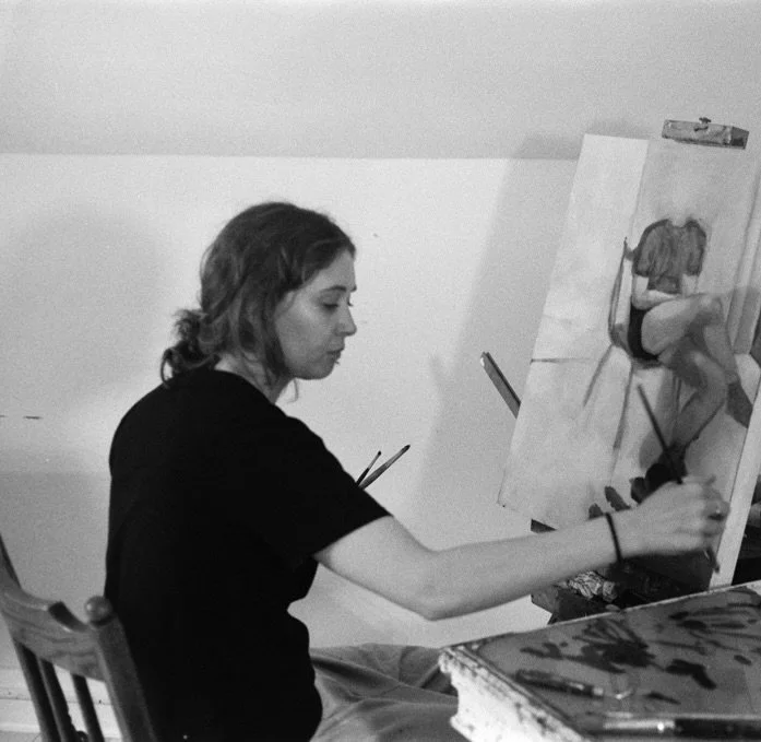 A woman seated in a chair is painting in her studio.