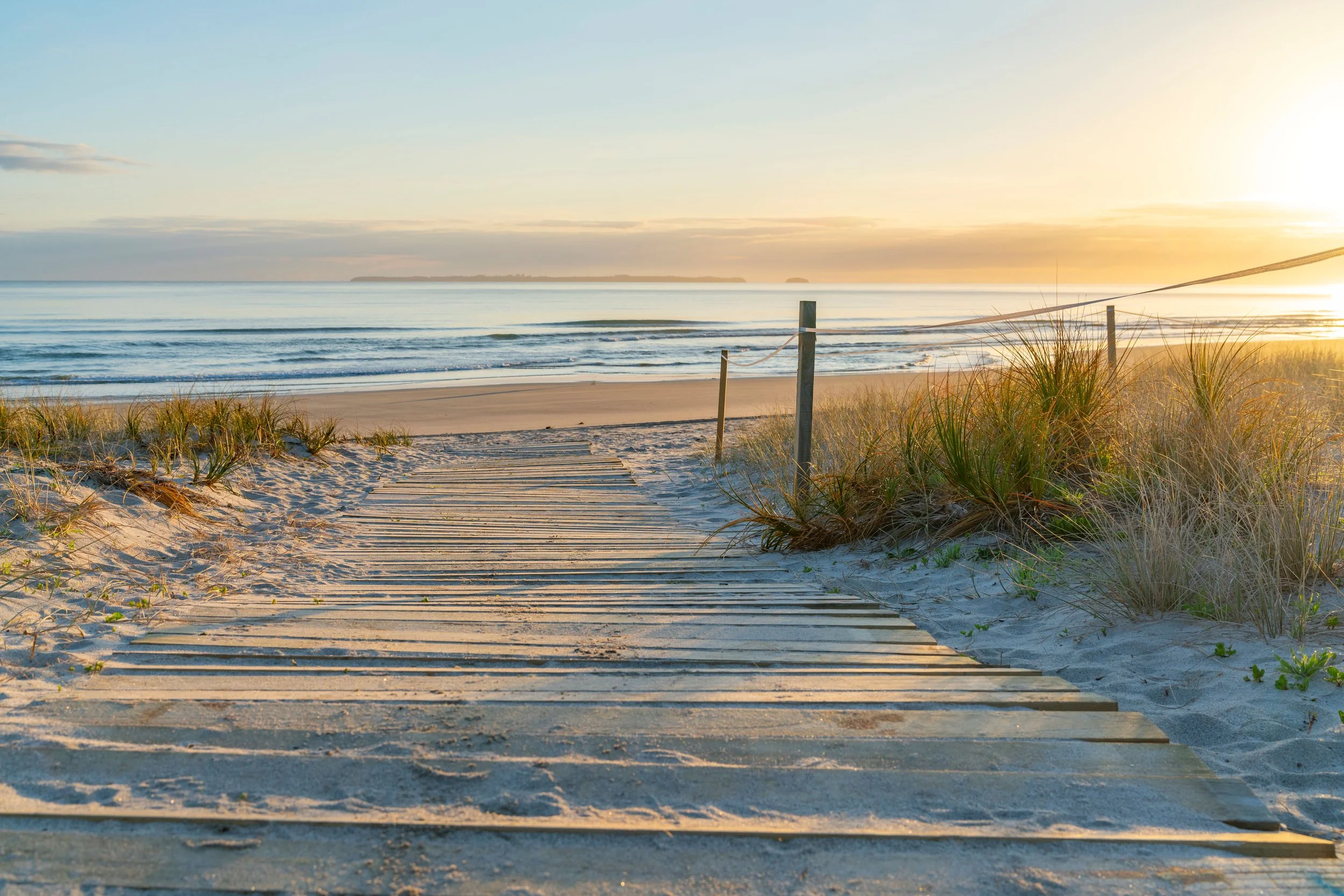 Sunset over a sandy beach with a wooden pathway, dune grass, and the ocean in the background.