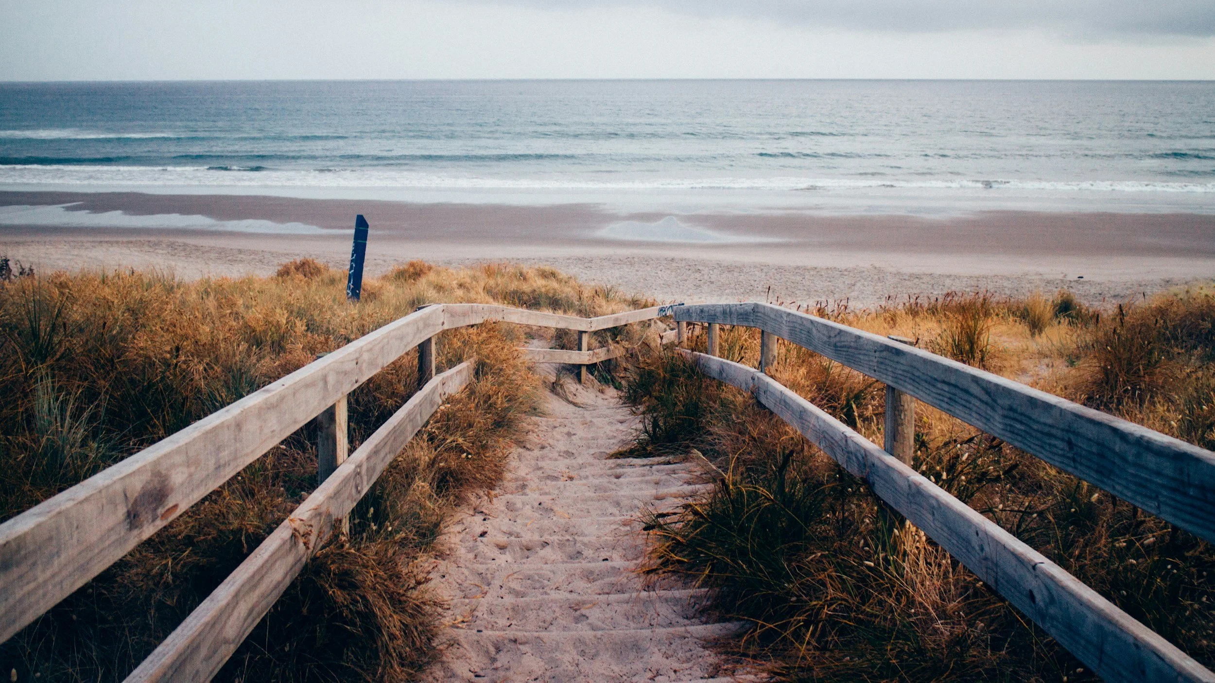 Wooden stairs leading down to a sandy beach with grassy dunes, ocean waves, and overcast sky in the distance.