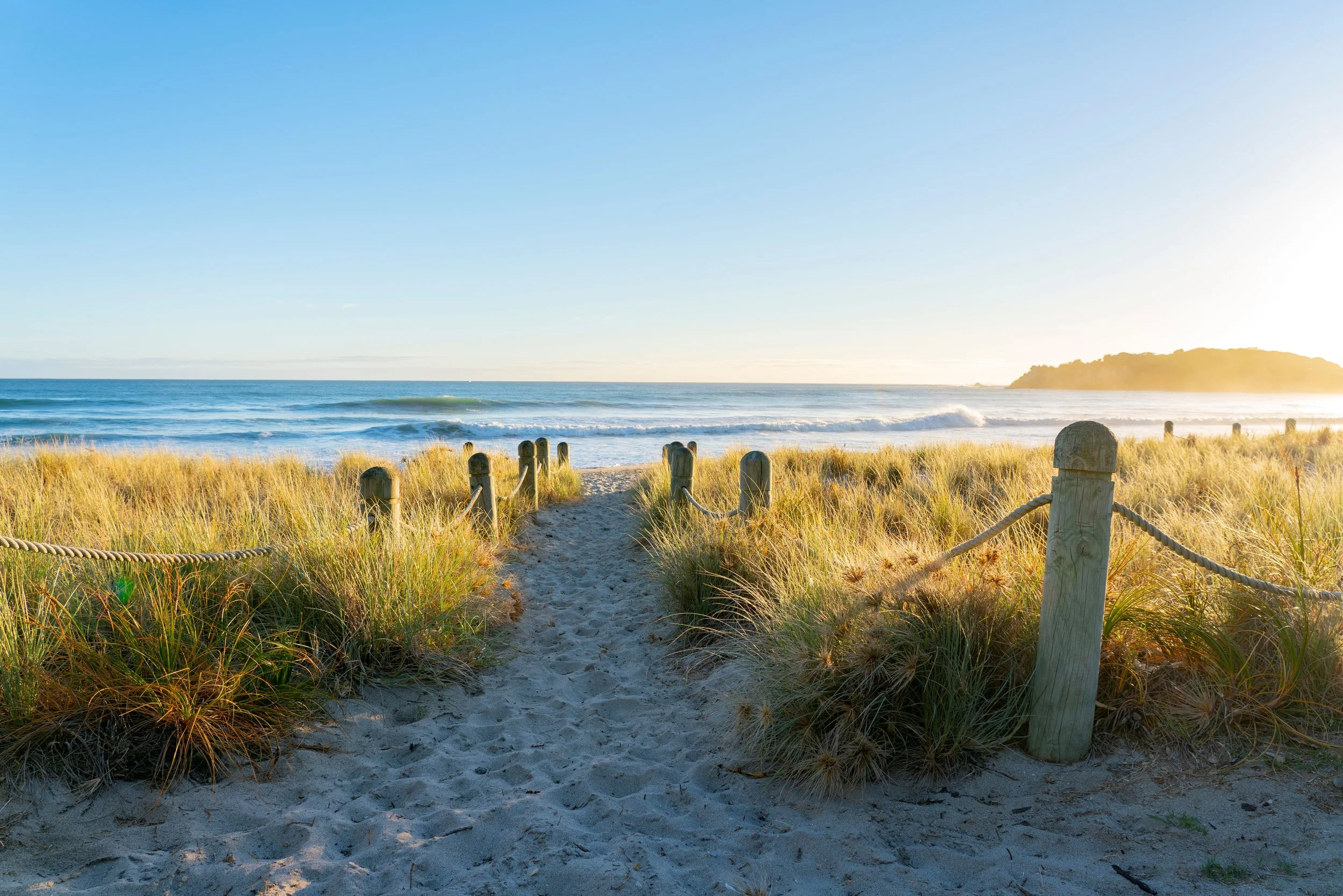 Path leading to the beach with sand dunes, grass, and ocean waves under a clear sky at sunset.