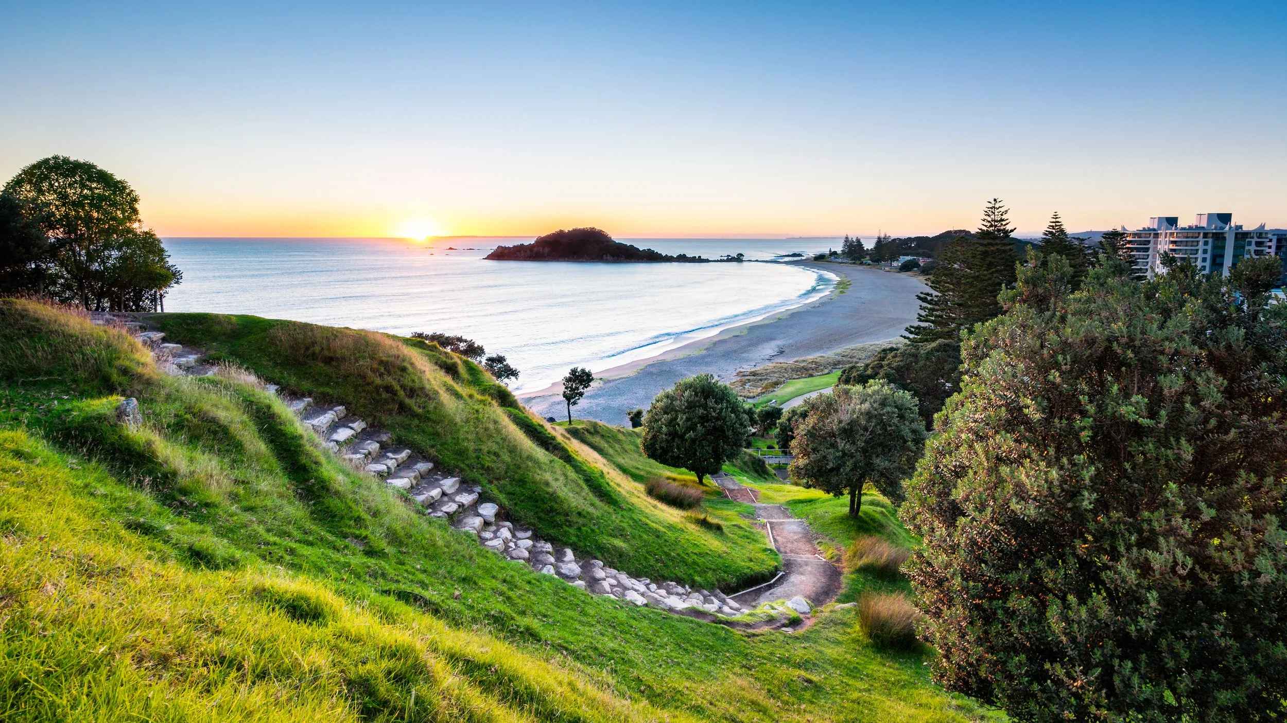 Sunset over a scenic coastal landscape with green grassy hills, trees, a pathway, and buildings nearby.