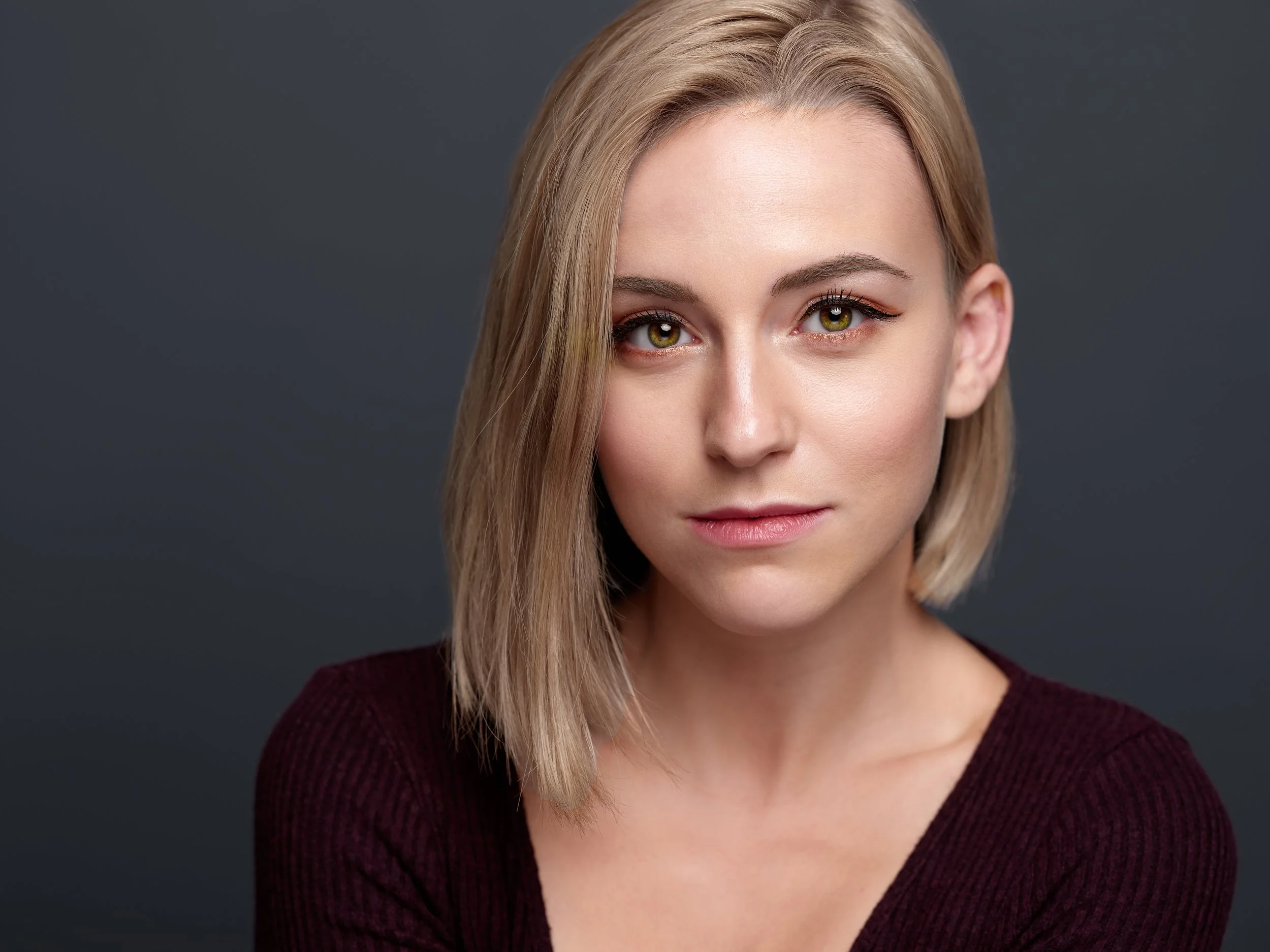 Close-up portrait of a young woman with fair skin, straight blonde hair, and hazel eyes, wearing a burgundy top against a dark gray background.