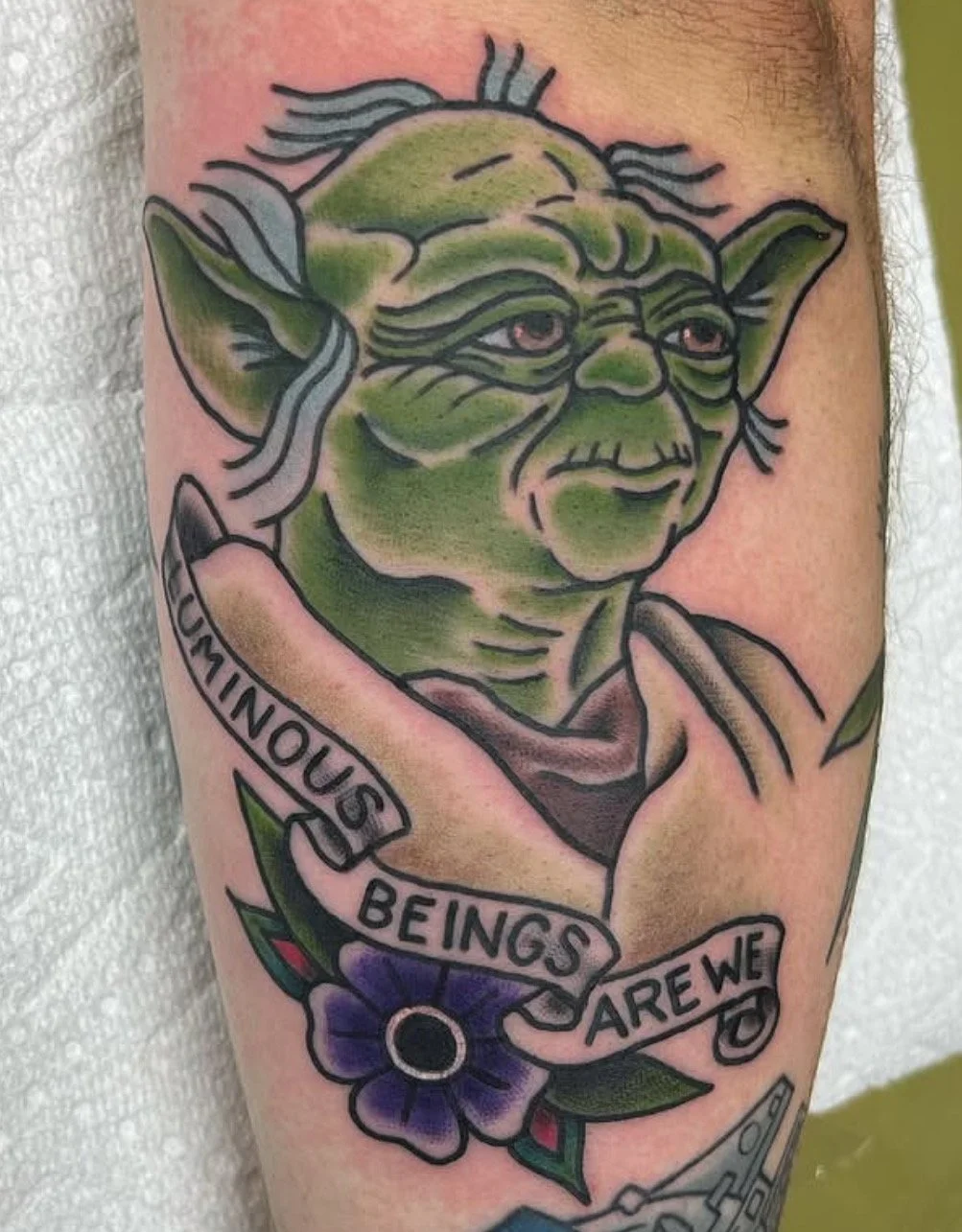 Tattoo of Yoda from Star Wars with a banner that reads 'Continuous Being's Are We' and a purple flower with green leaves.