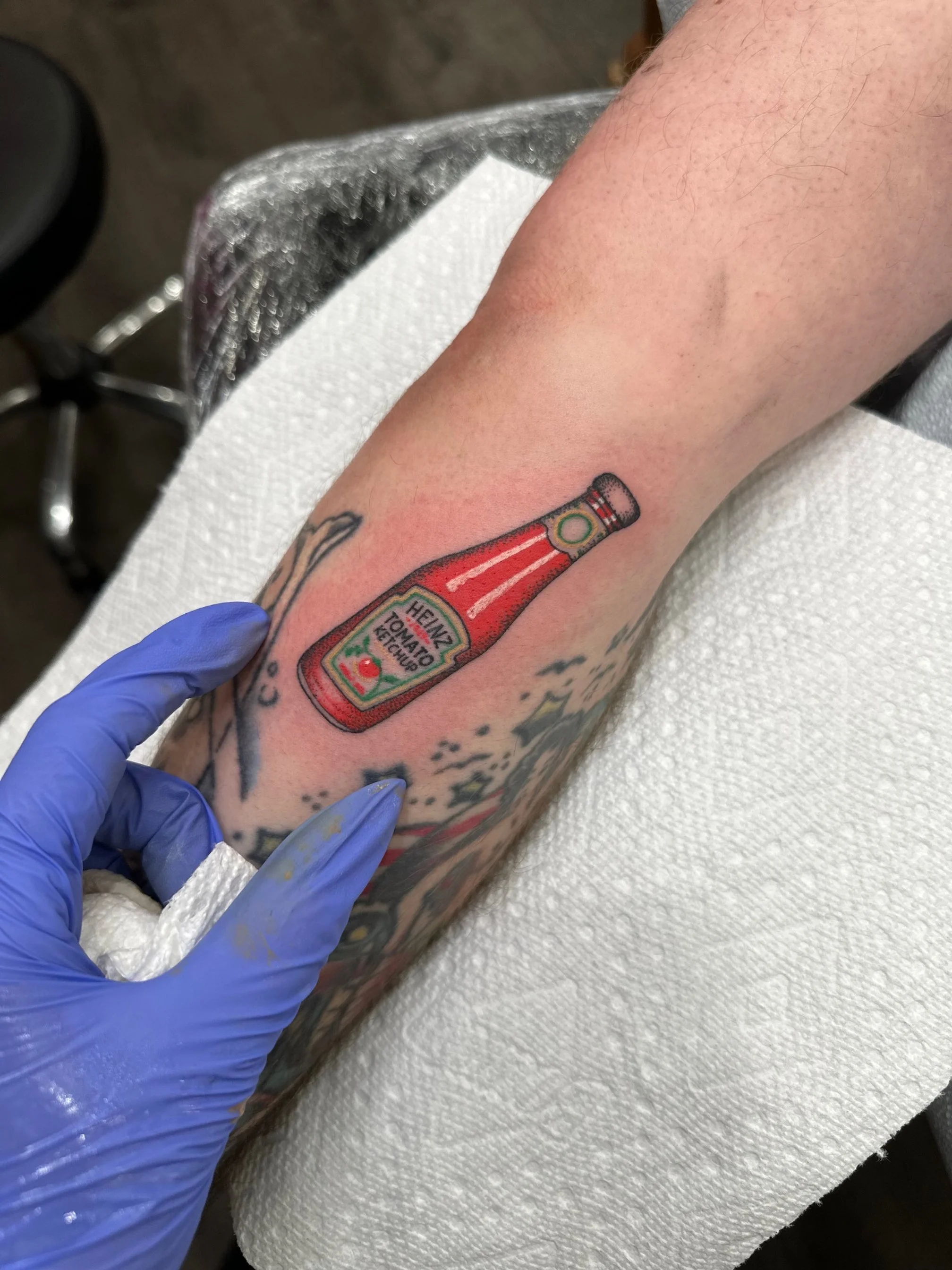 Tattoo of a ketchup bottle on a person's forearm in a tattoo studio.