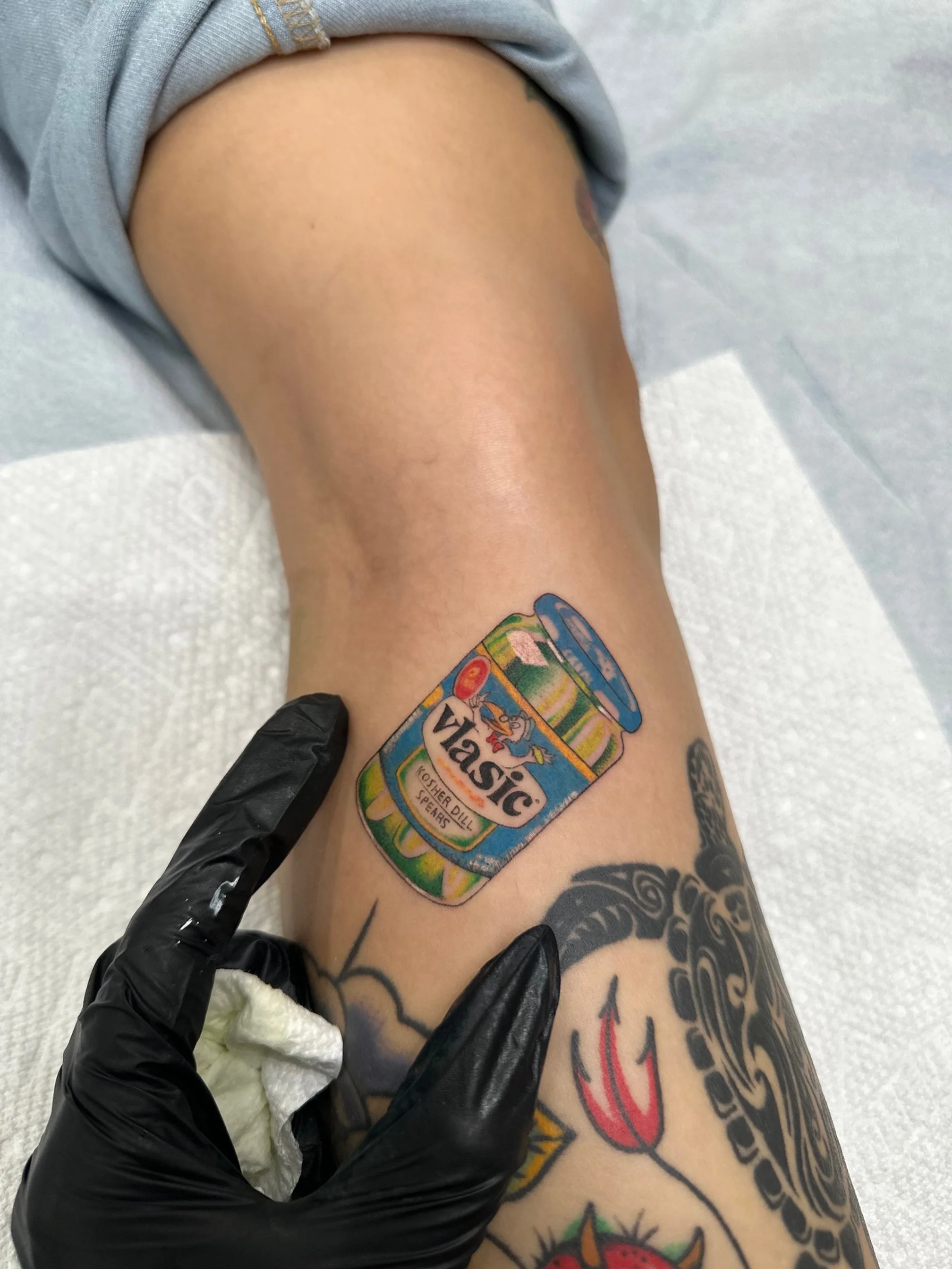Tattoo of a large colorful jar labeled "Vlassic" with a cartoon duck, on a person's leg alongside other tattoos, while a gloved hand holds a paper towel nearby.