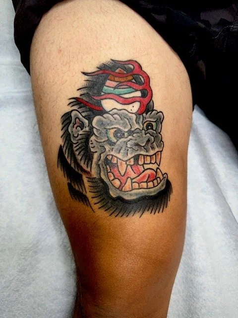 Tattoo of a stylized, fierce lion's head with an open mouth, featuring an abstract, wavy colorful element above it on a person's thigh.