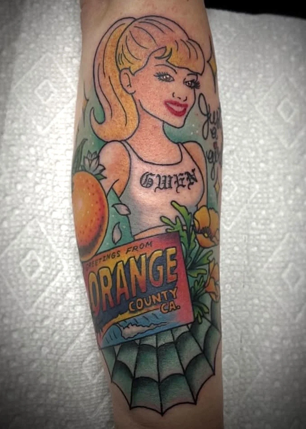 Colorful tattoo of a smiling woman with blonde hair, a tattoo on her chest, a strawberry, a spider web, and a sign that reads "Greetings from Orange County, CA."