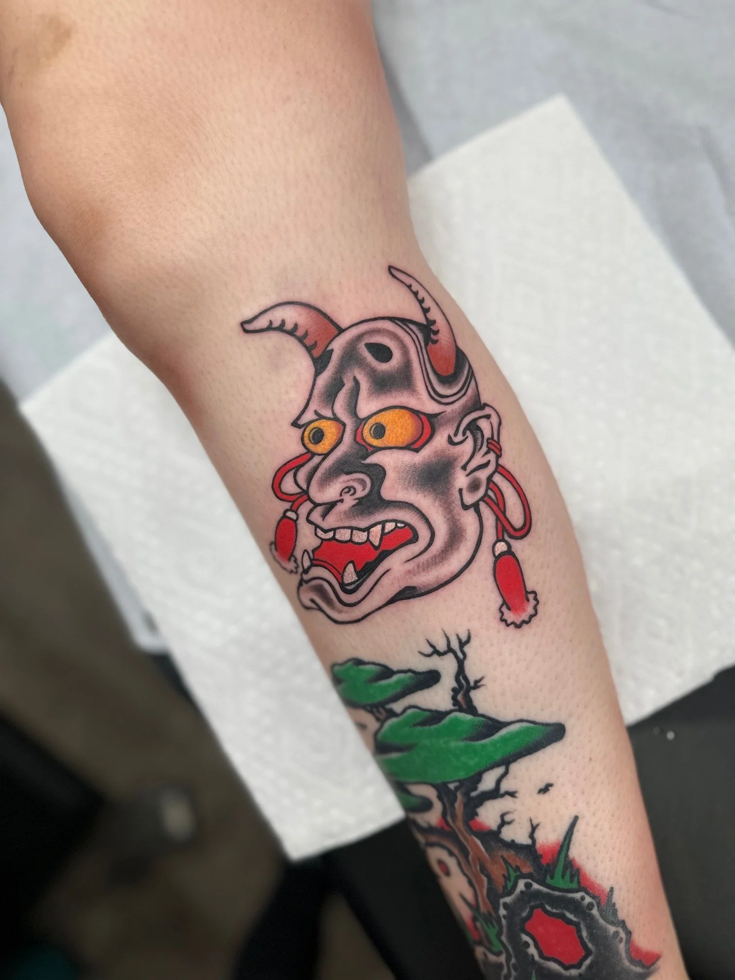Colorful tattoo of a demonic face with horns, yellow eyes, and red earrings on a person's arm