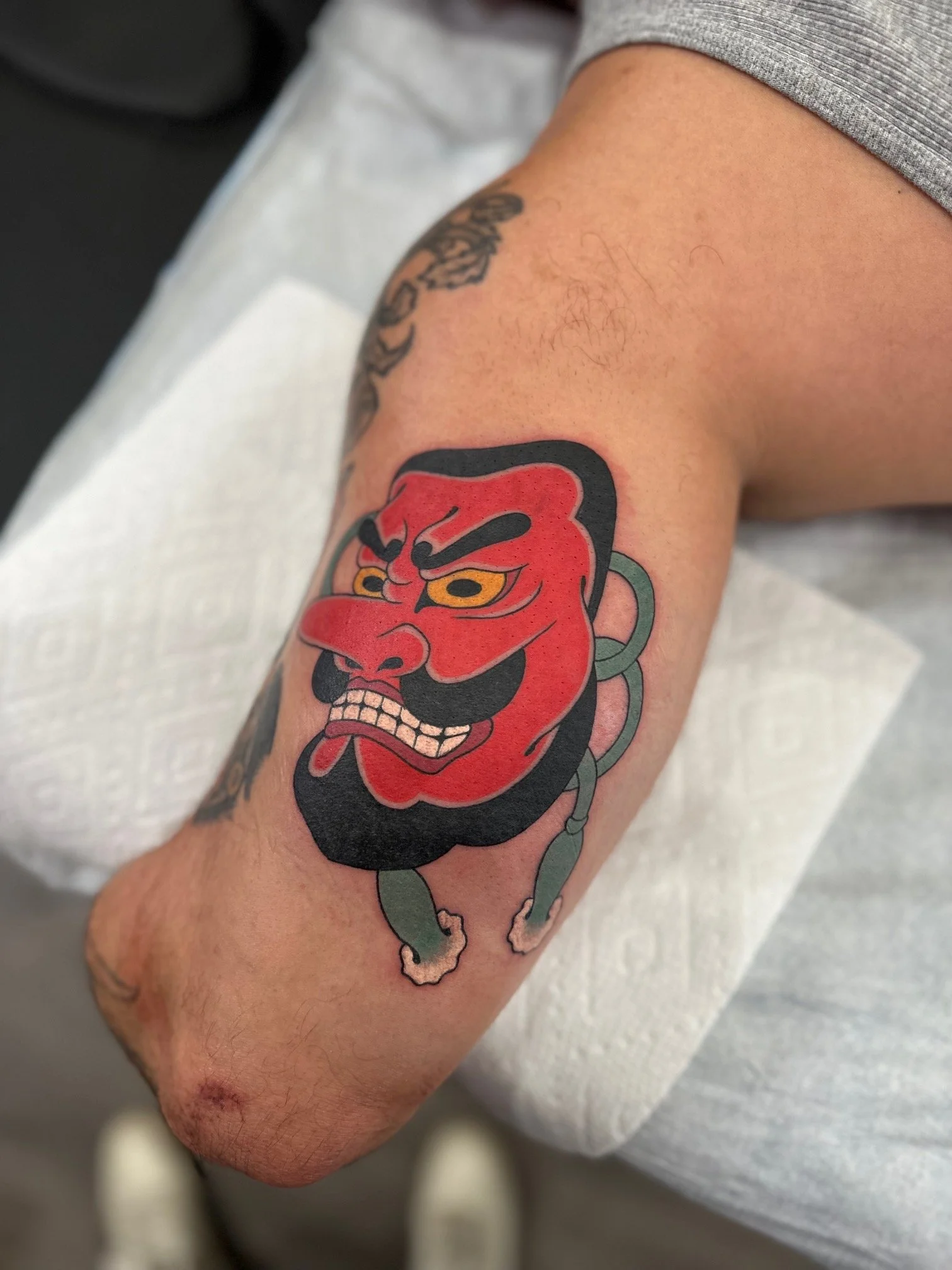 Tattoo of a red-faced devil with black hair, eyebrows, and beard, yellow eyes, gritted teeth, and a menacing expression on a person's upper arm.