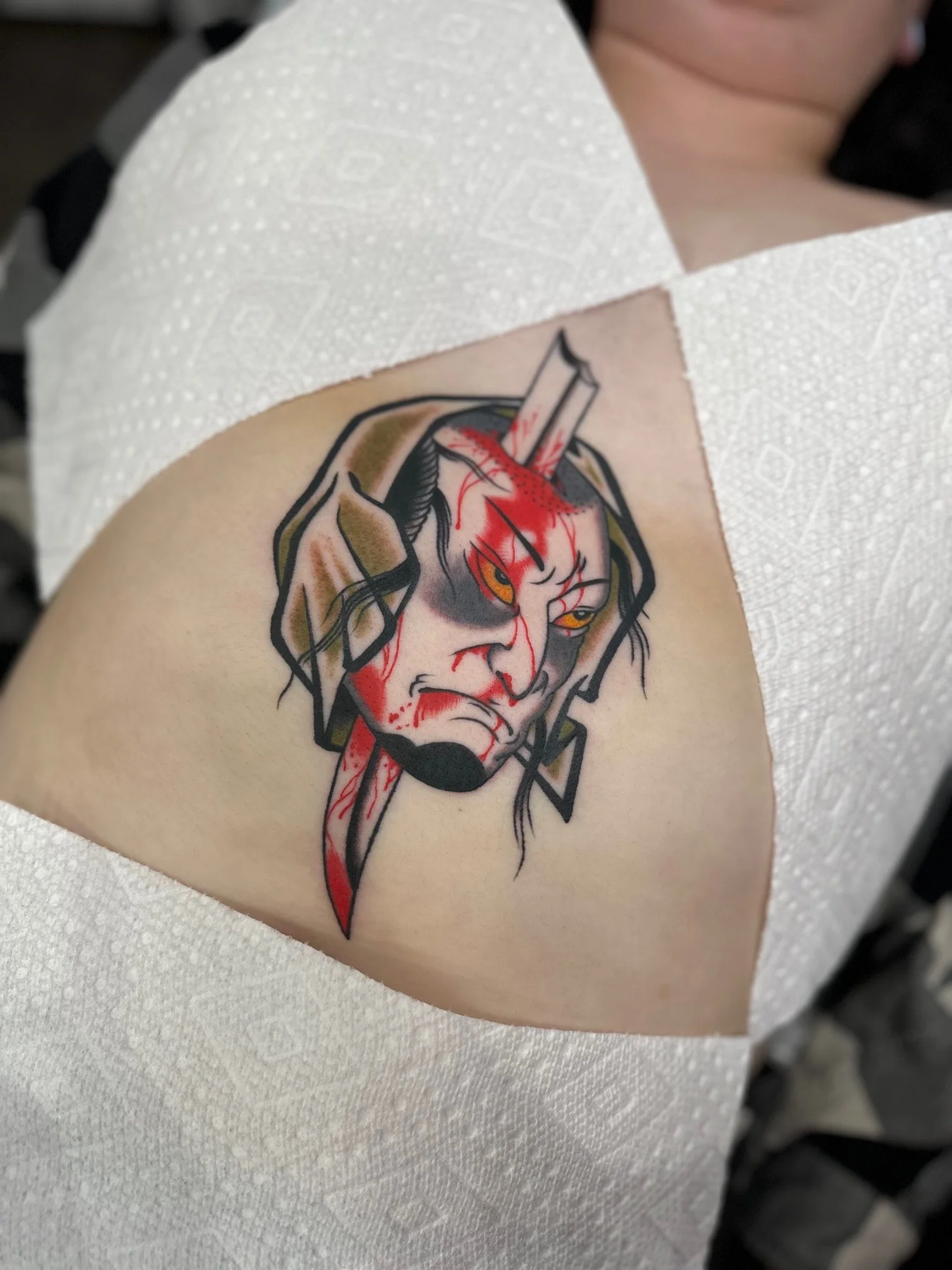 Tattoo of a fierce, angry Oni or demon mask with blood streaks, black hair, and a prominent dagger piercing through its forehead.
