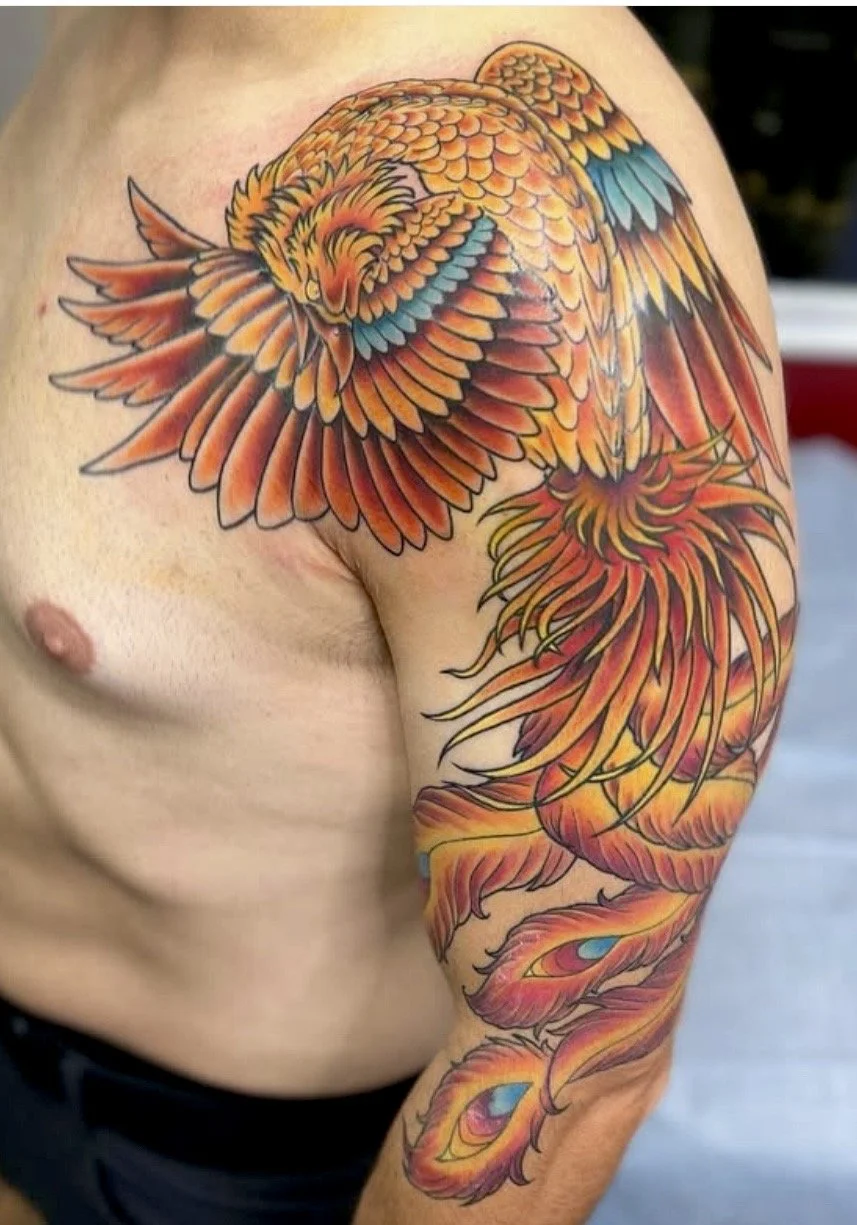 Colorful tattoo of a phoenix with orange, red, yellow, and blue feathers on a person's upper arm and shoulder.