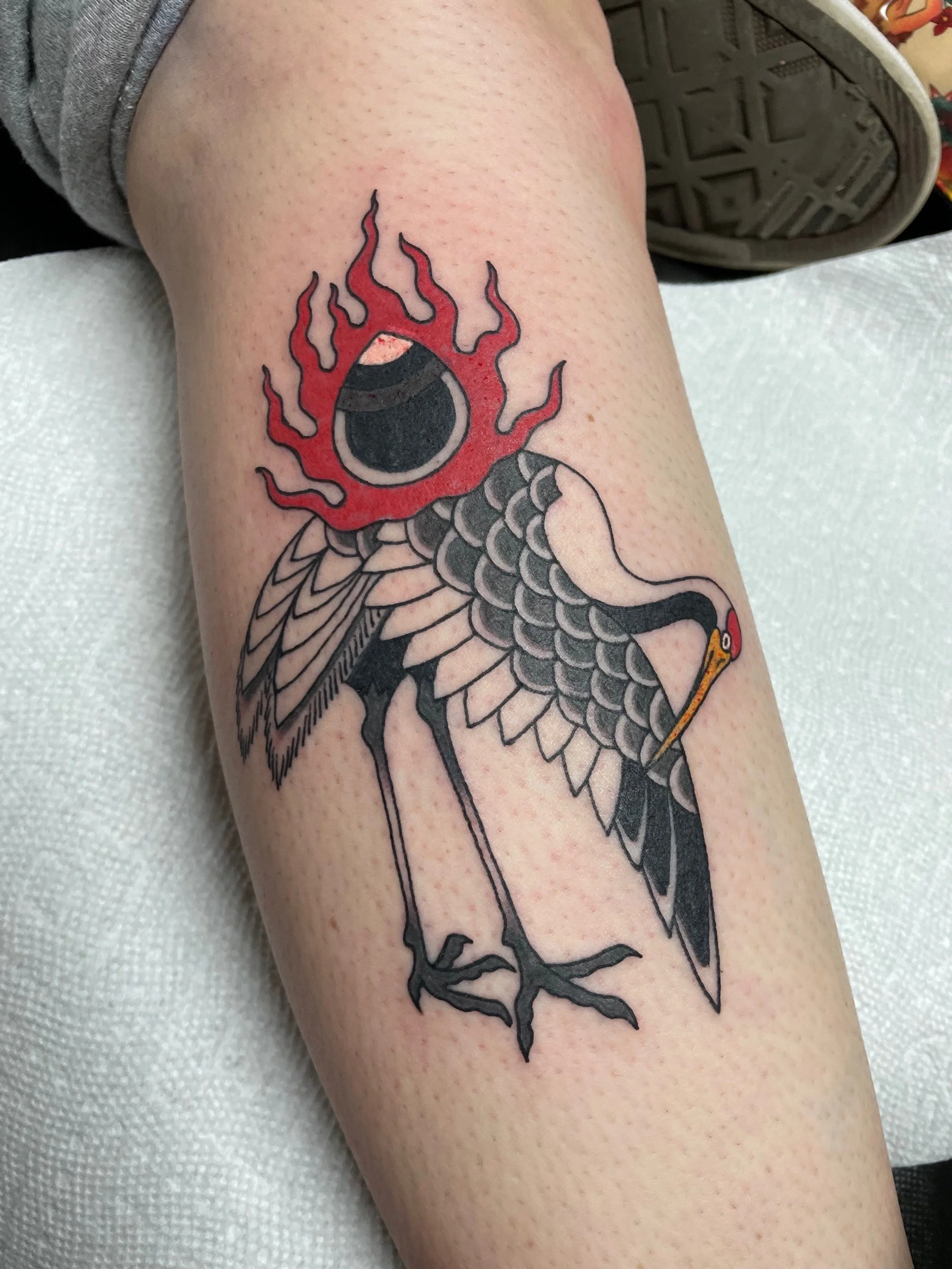 Tattoo of a crane with a large eye surrounded by red flames on its head and black details, depicted in a traditional style.