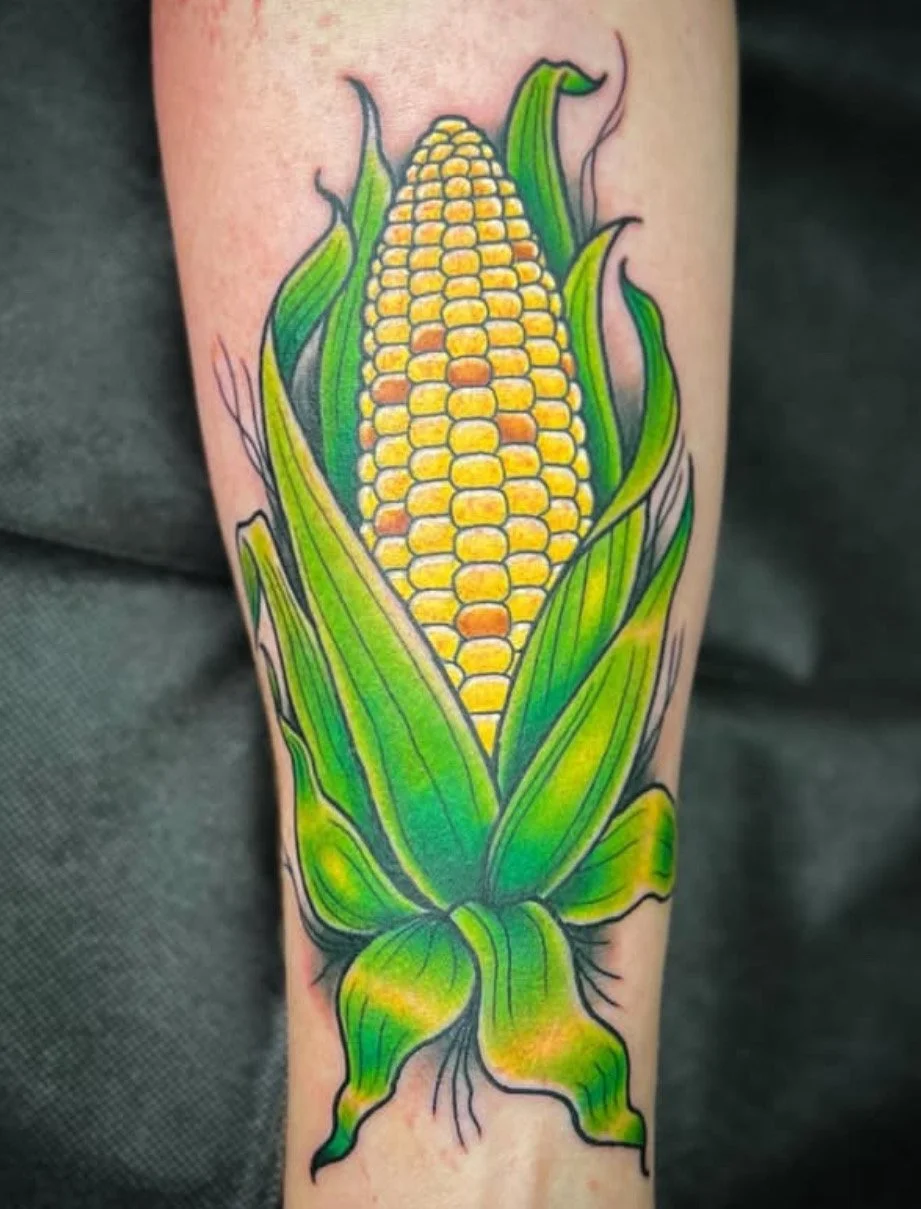 Colorful tattoo of a yellow corn cob with green leaves on a person's arm.