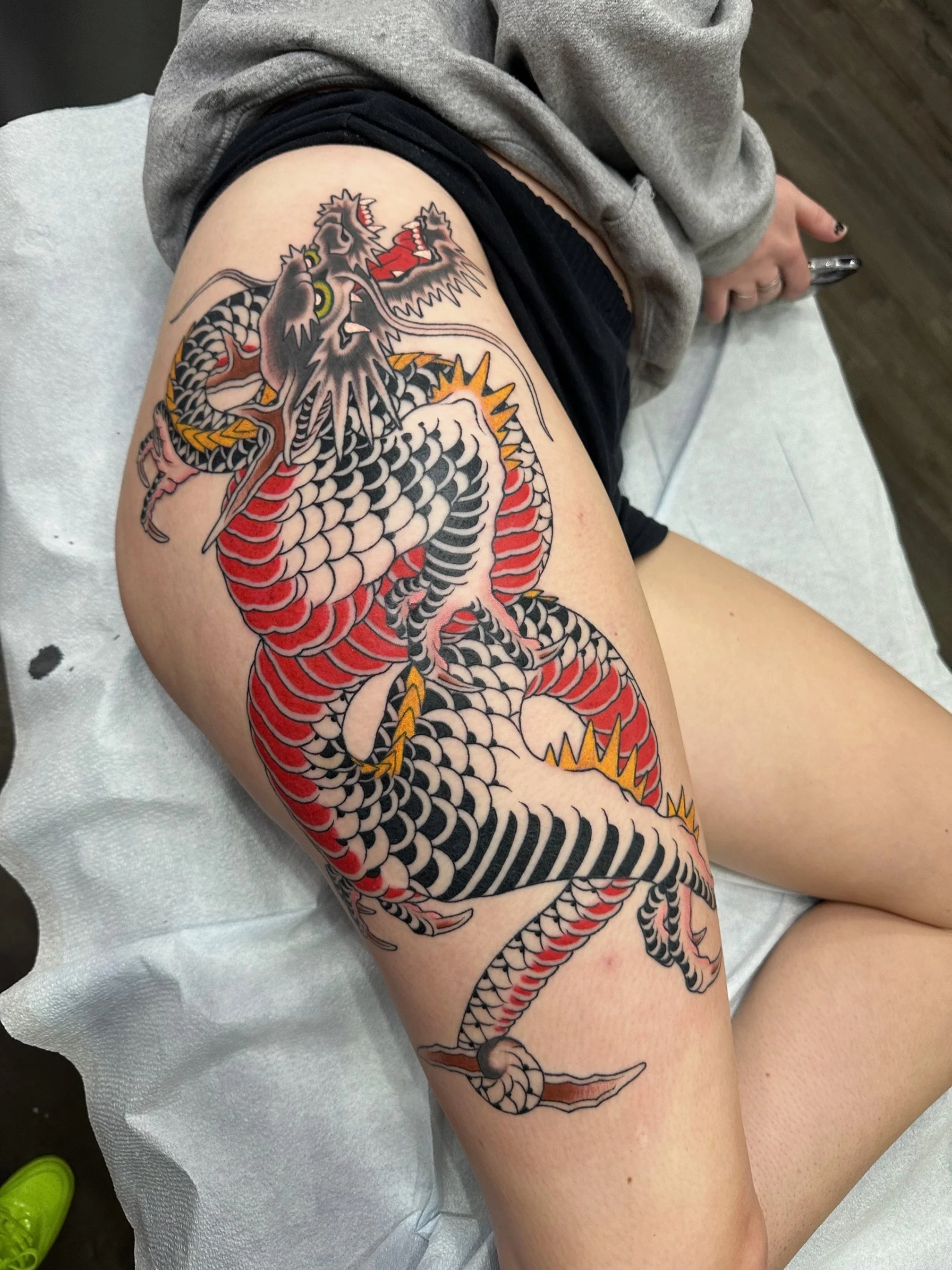 A detailed tattoo of a traditional Japanese dragon on a person's thigh, featuring black, red, white, and yellow colors with intricate scales and fierce facial features.