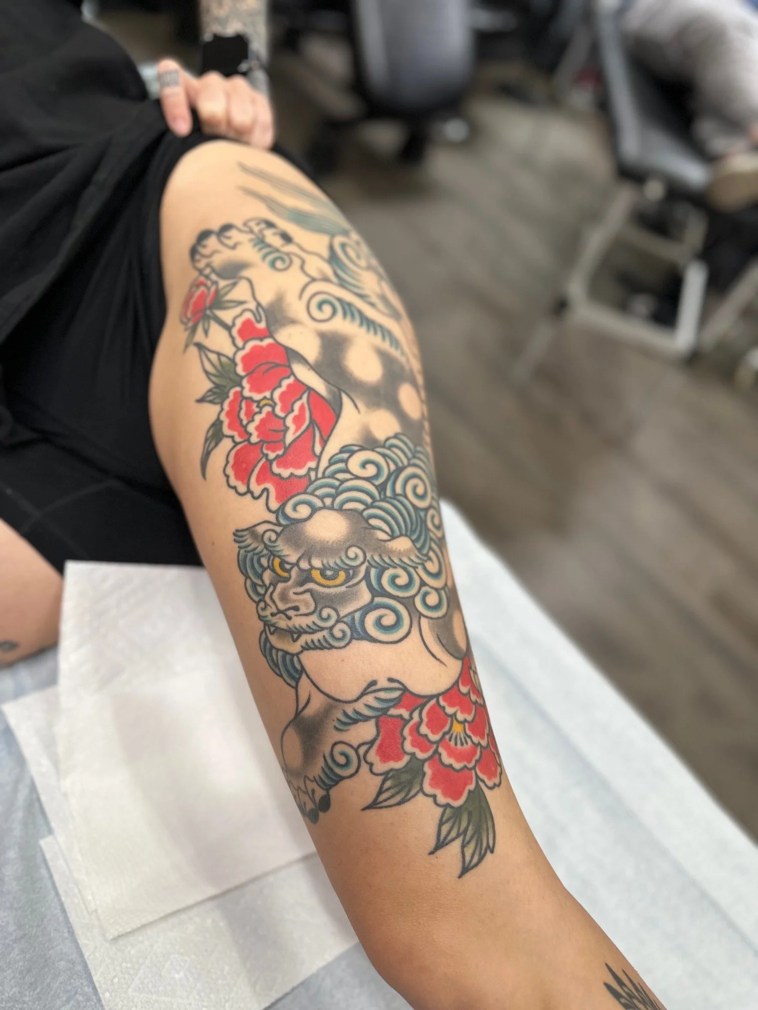A colorful tattoo of a traditional Japanese style lion or tiger with blue clouds and red flowers on a person's upper arm.