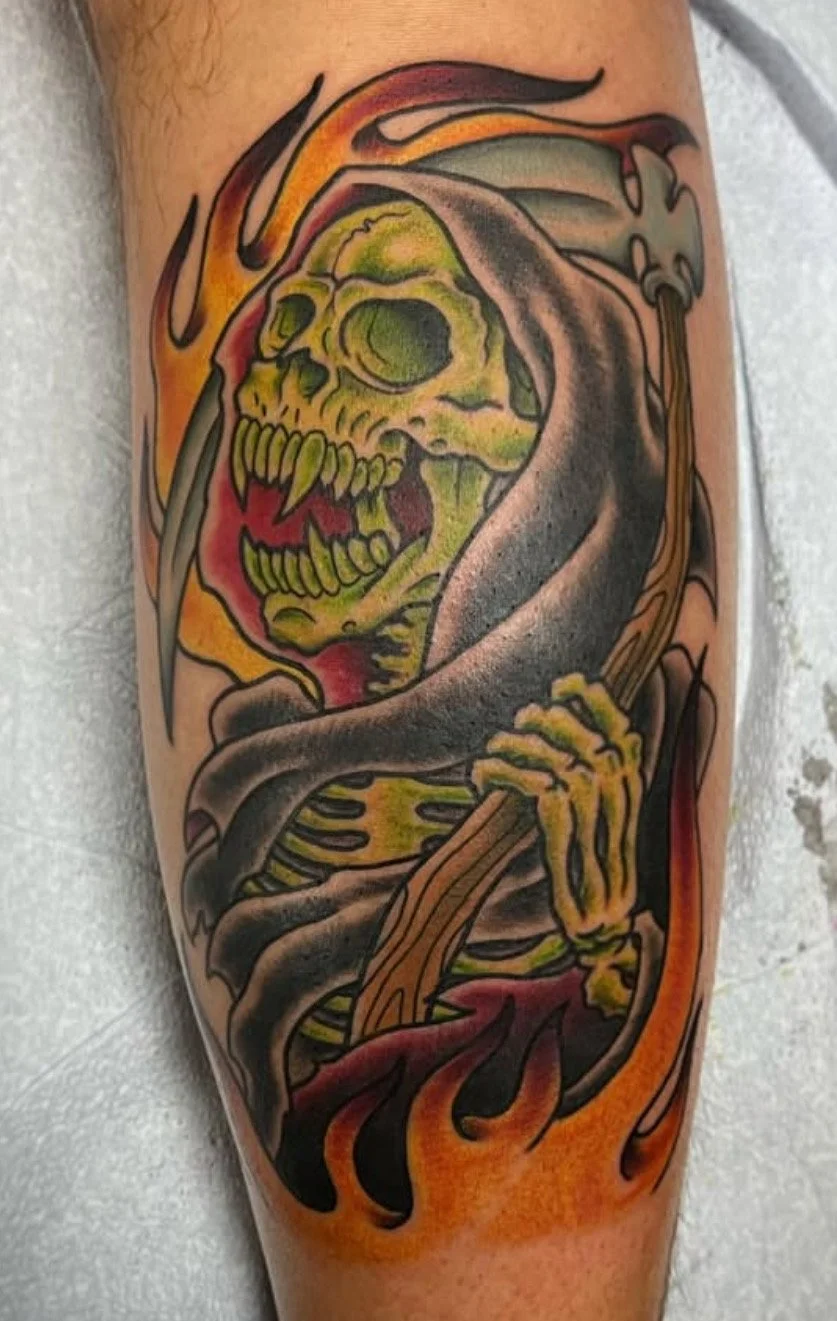 Tattoo of a skeleton with a green skull and glowing eyes, cloaked in dark gray and black fabric, holding a staff, surrounded by flames.