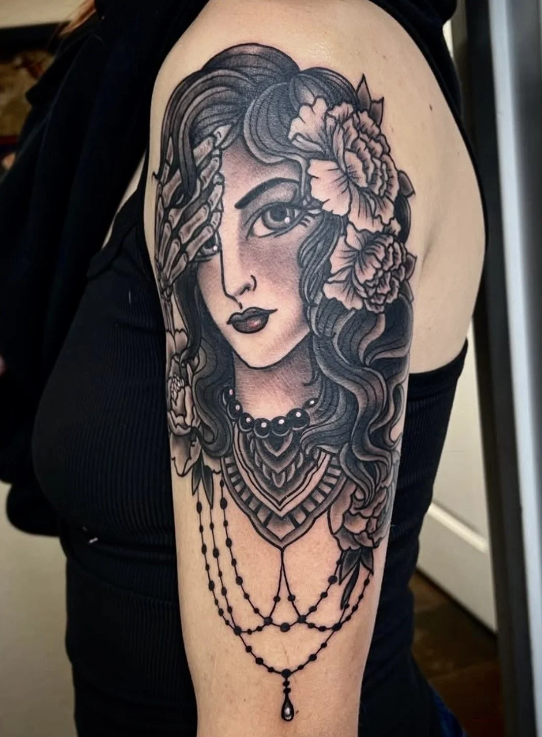 Black and gray tattoo of a woman with long wavy hair, adorned with flowers and necklaces, covering the upper arm and shoulder.