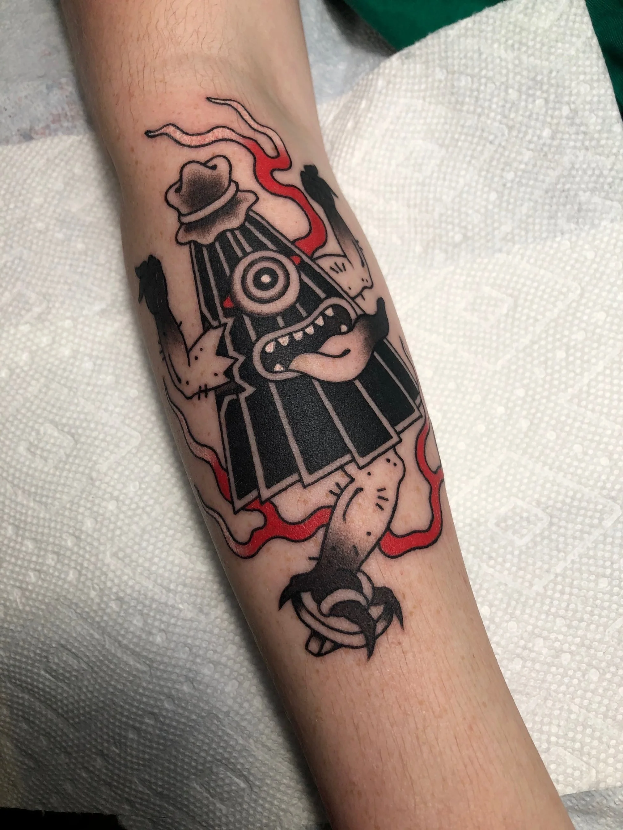 Tattoo of a cartoonish monster with a single eye, wearing a hat, with tentacle-like arms and a paw at the bottom, outlined in black with some red highlights.