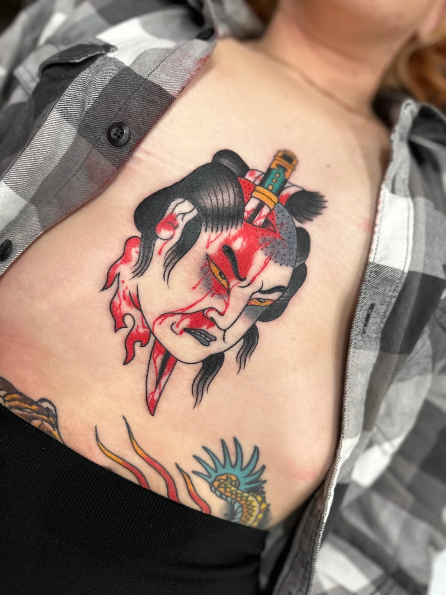 Tattoo of a fierce woman with traditional ninja or samurai warrior makeup and hairstyle, with a dagger and a small sword on her forehead, and a snake below her chin.