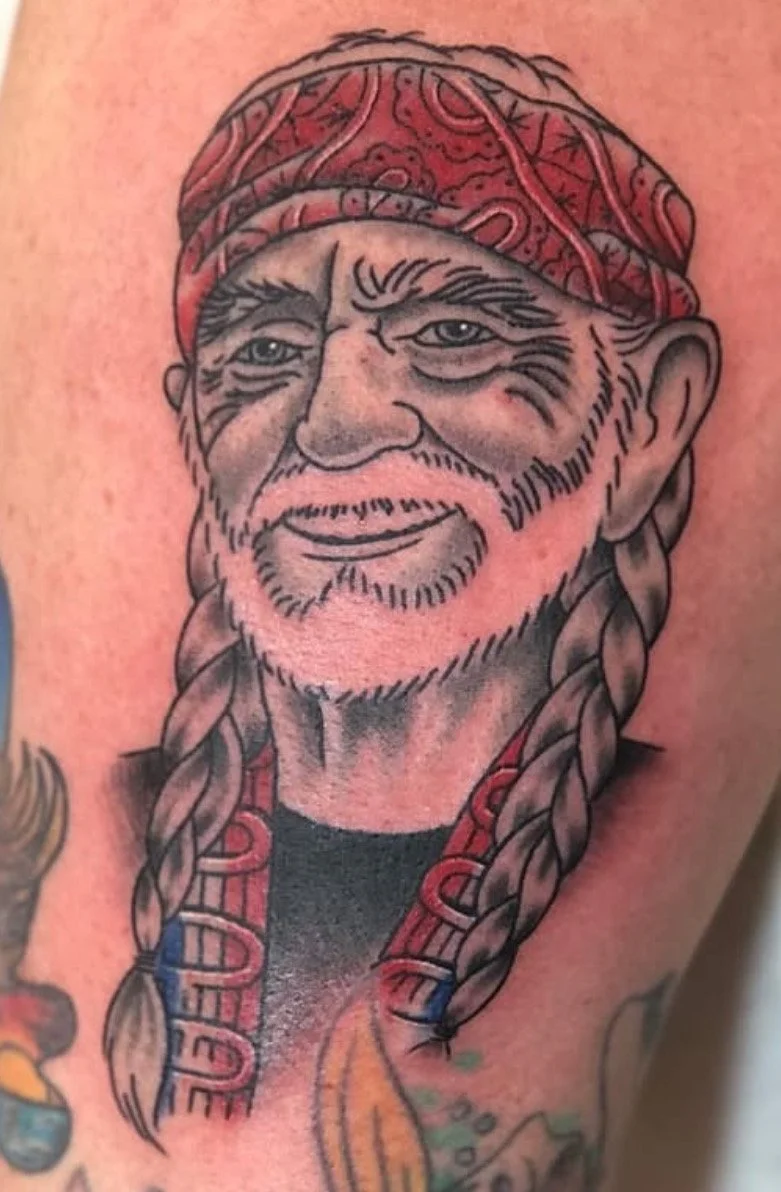 Tattoo of a smiling older man with a beard and mustache, wearing a red bandana and a black shirt.