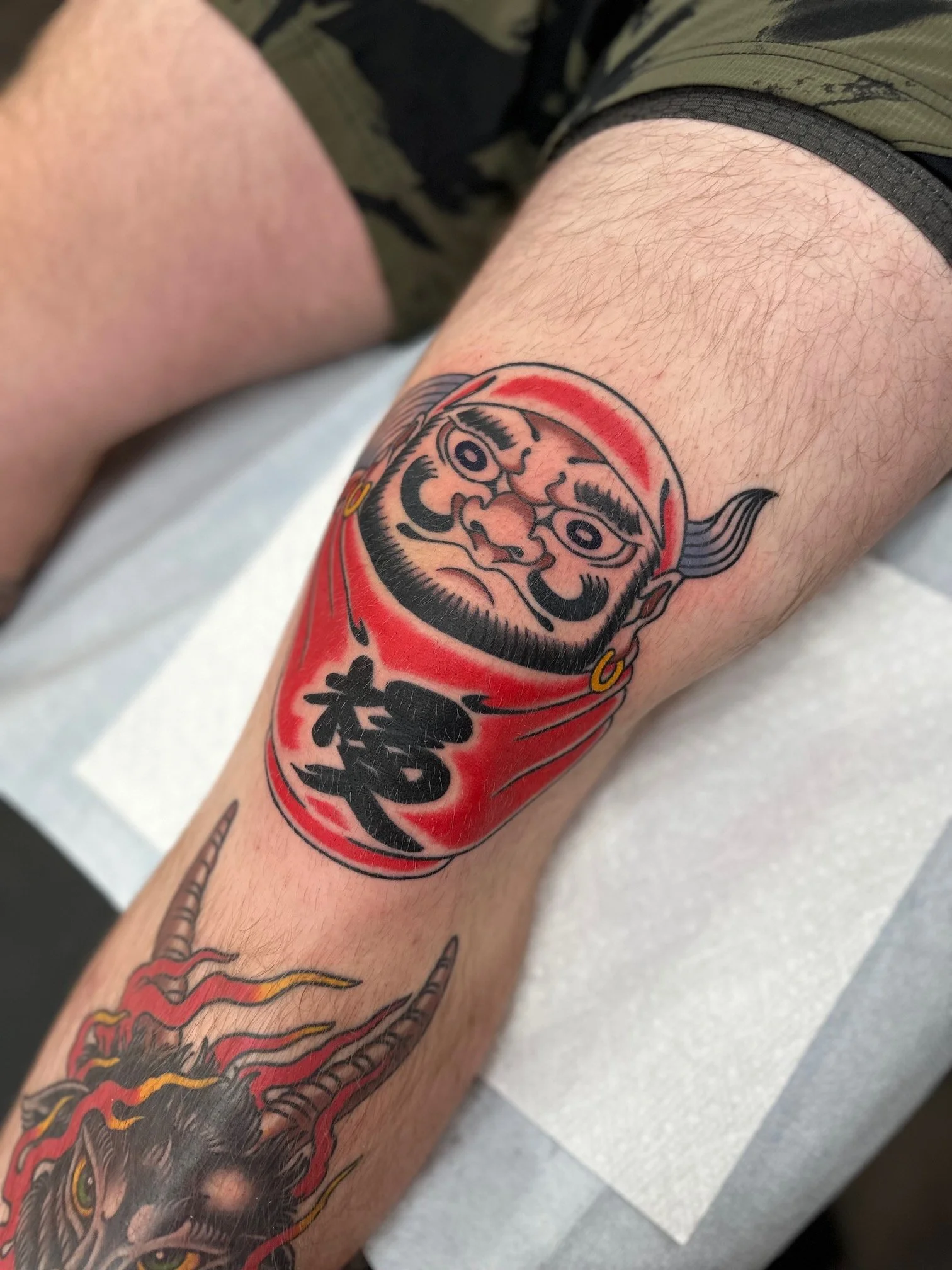 Tattoo of a Daruma doll with a fierce facial expression on a person's calf, with the doll's face featuring bold black outlines, red accents, and Japanese characters.