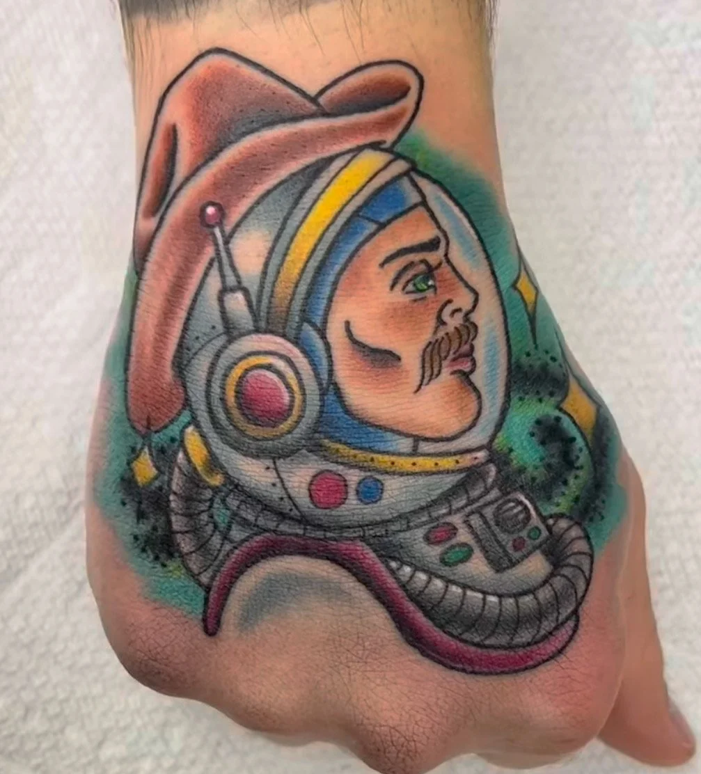 Colorful tattoo of a man with a mustache wearing an astronaut helmet and suit, with a background of green and black ink.