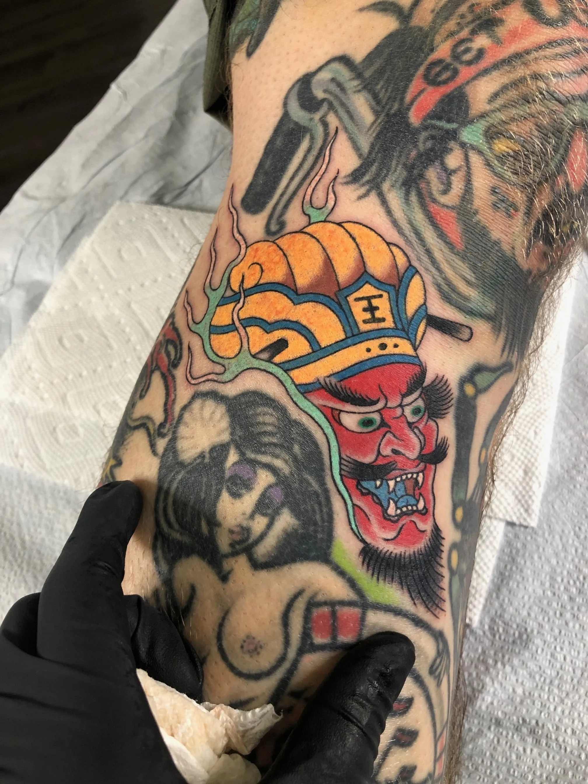 Colorful tattoo of a demon with a red face, black hair, mustache, wearing a blue and orange hat with Chinese characters, and a woman with dark hair, on a person's arm.