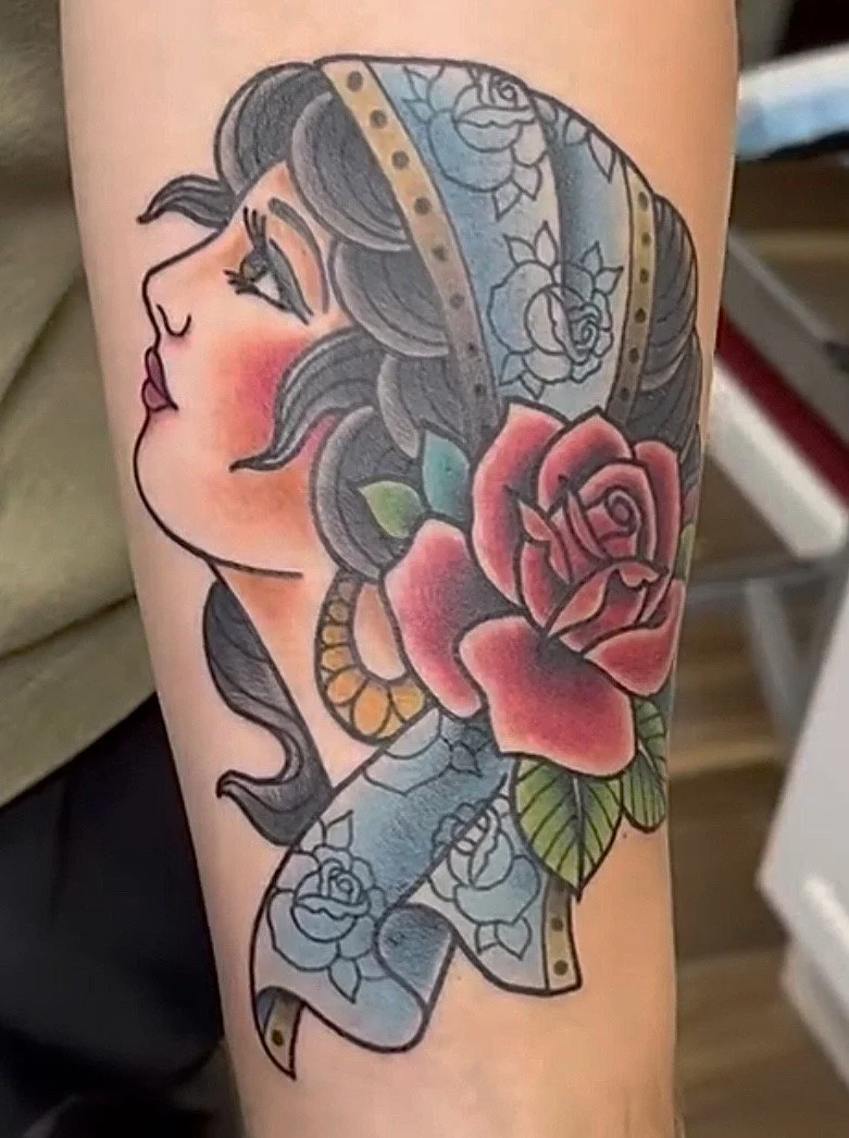 Tattoo of a woman's face in profile with flowing hair, a large pink rose, green leaves, and a blue floral sash.