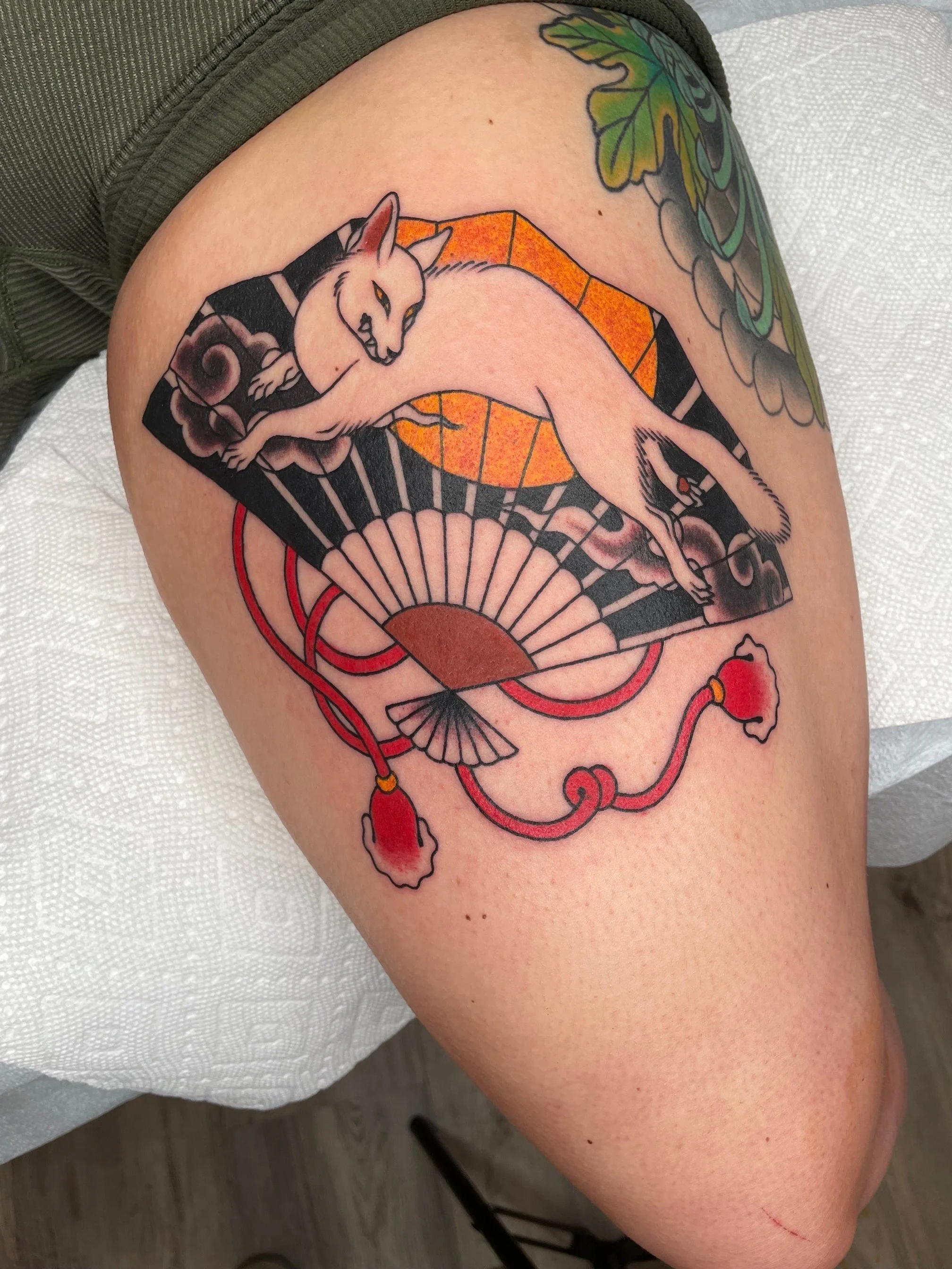 Tattoo of a reclining white fox cat hybrid with a stylized background including a fan and decorative elements.