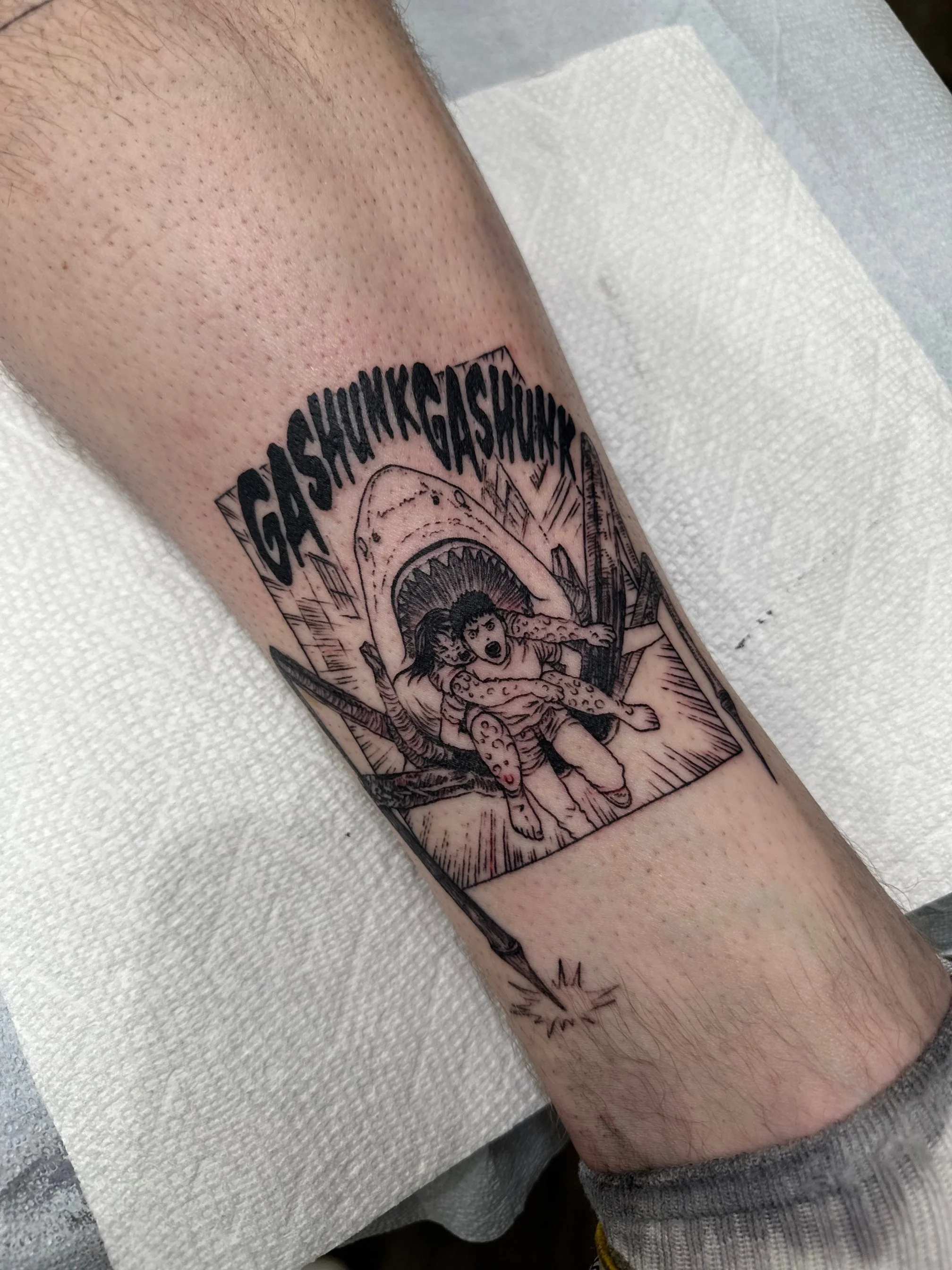 A tattoo featuring a comic-style illustration of a person with wide eyes, seemingly frightened, being attacked by a large creature with a gaping mouth, surrounded by jagged mountains and the word 'GASSHHHHHKASKSHKH' at the top.