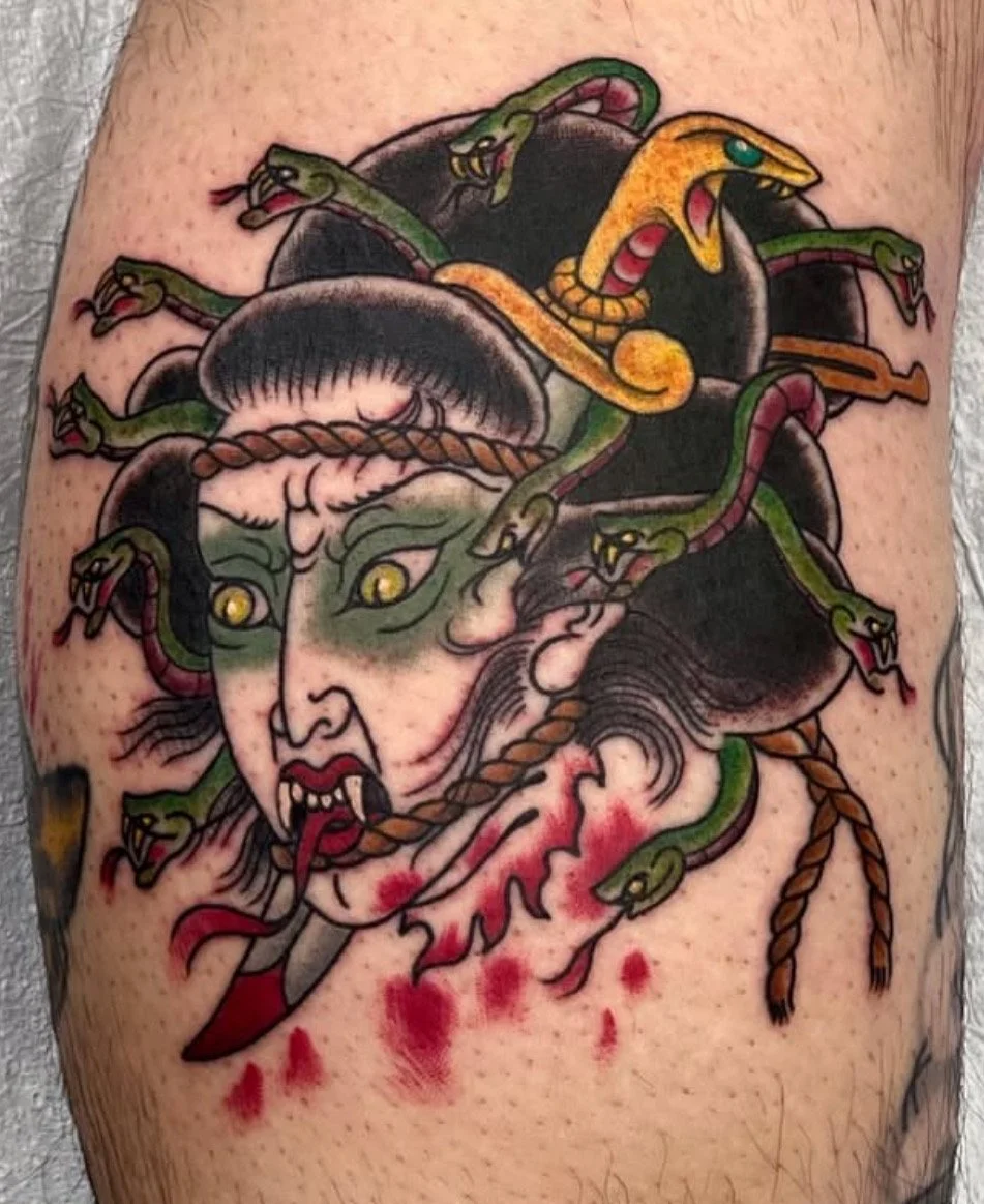 Tattoo of a woman with a fierce expression, green eyes, dark hair, and red lips surrounded by green and yellow snakes with open mouths, some with visible fangs, and a blood-like splatter at the bottom.