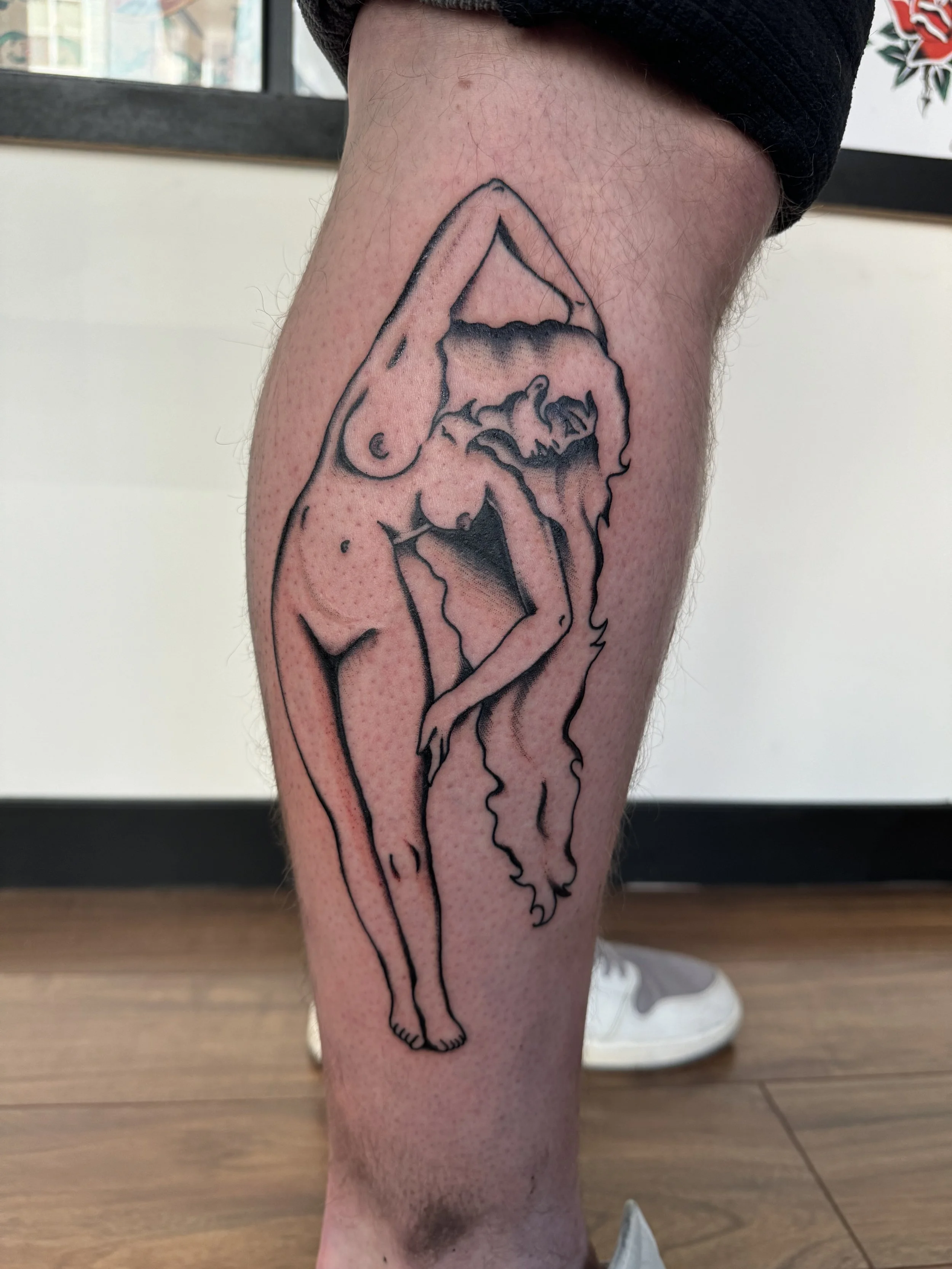 Tattoo of a stylized nude woman with long hair, bent forward, outlined in black ink on a person's calf.