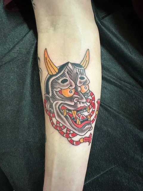 Tattoo of a traditional Japanese dragon or demon mask on a person's forearm, featuring horns, a fierce expression, and red and gray colors.