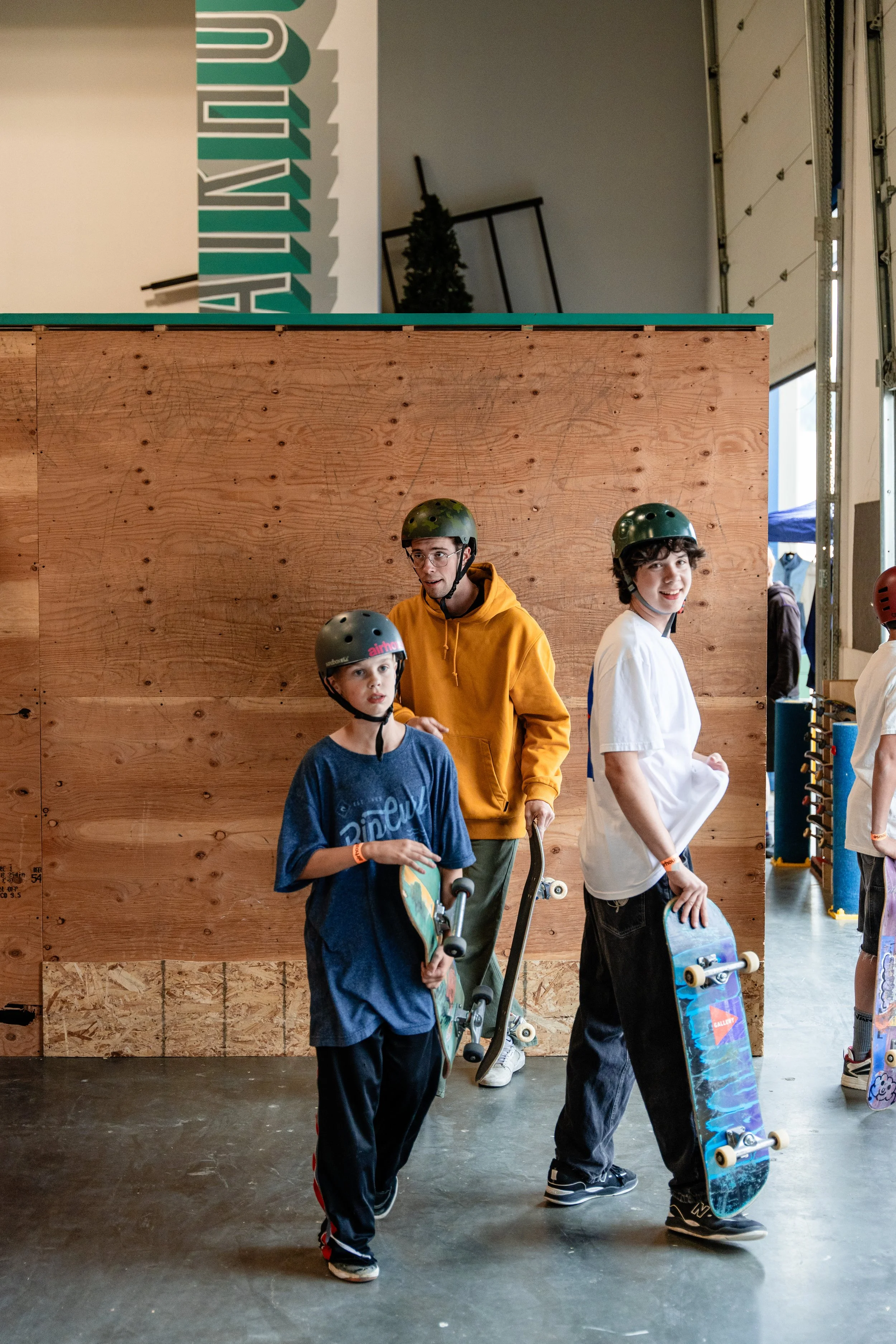 Three young boys wearing helmets and holding skateboards inside skate park with wooden ramp wall and open garage door in background.