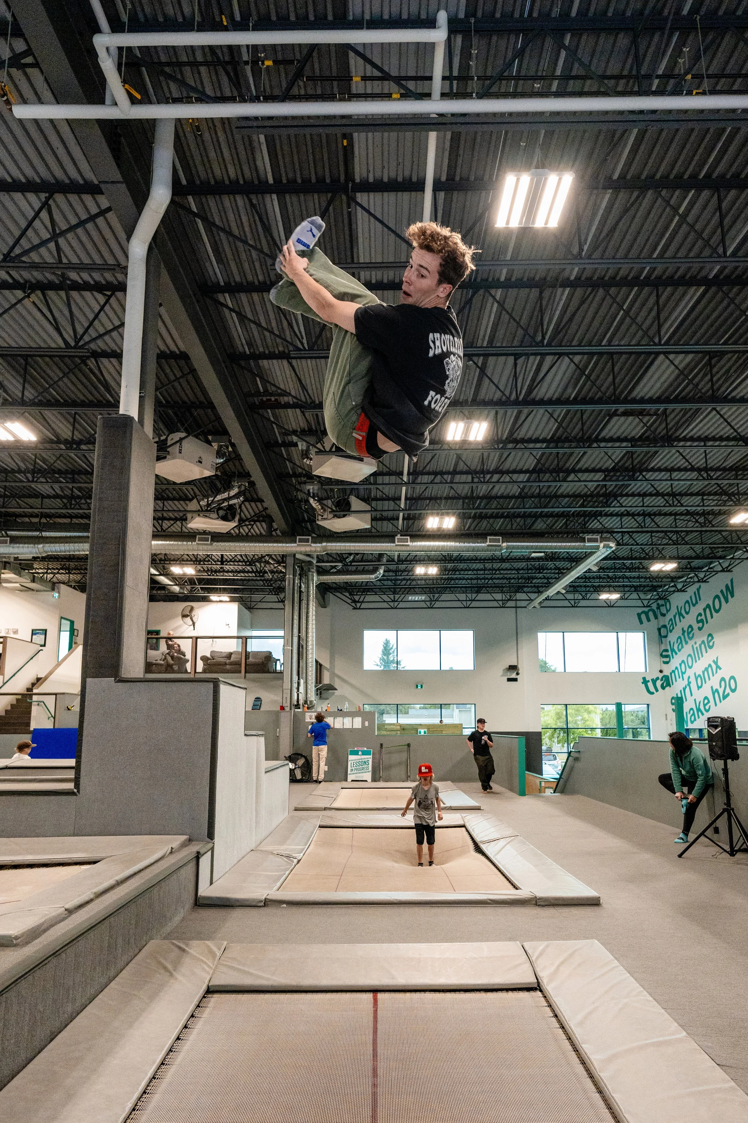 A person mid-air flipping at an indoor trampoline park with other people jumping and watching below.