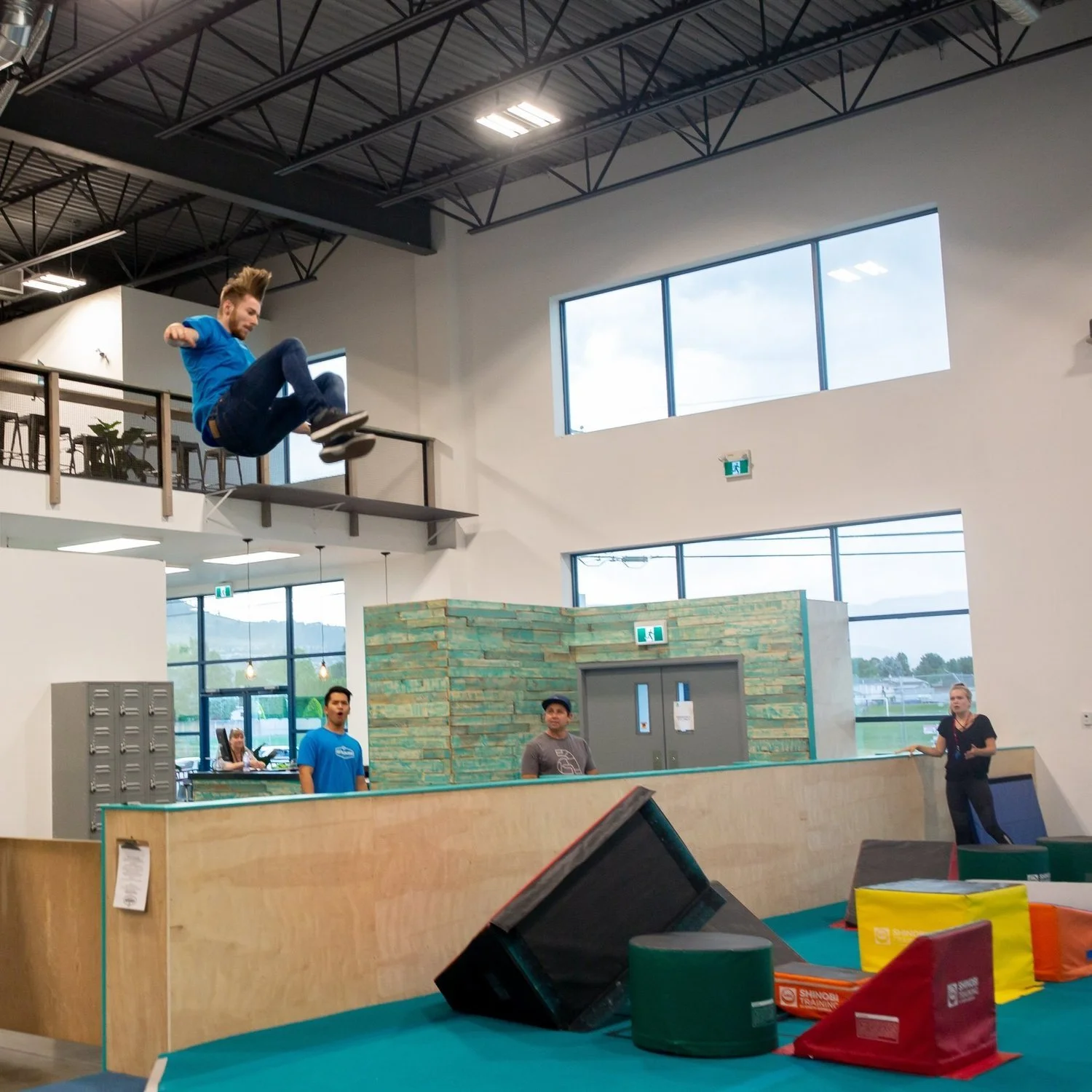 An indoor parkour training facility where a man is mid-air, performing a parkour move across a ledge or railing, while three other individuals watch from below.