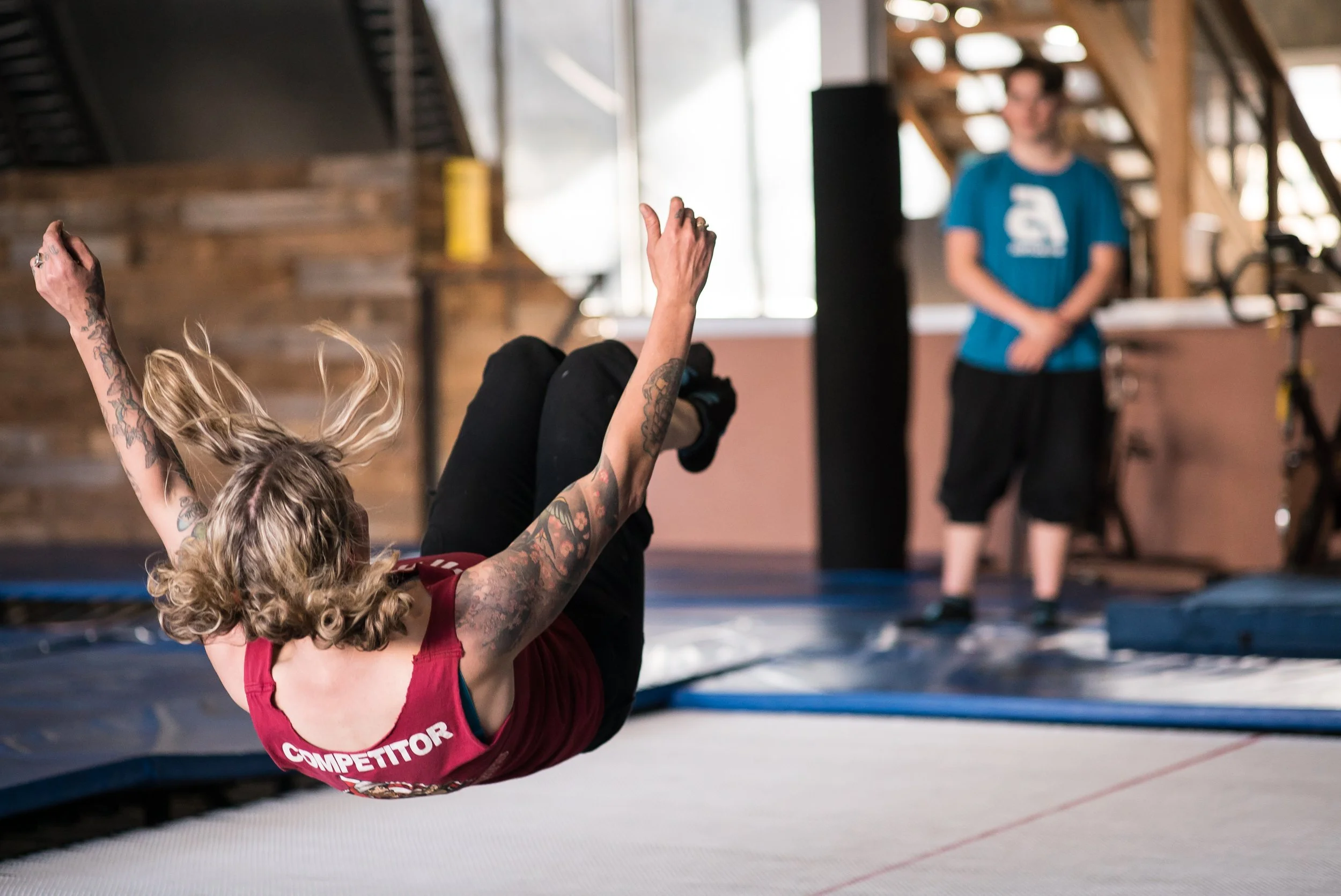 A woman with tattoos wearing a red shirt that says 'COMPETITOR' on the back, falling onto a trampoline in an indoor gym while a person in a blue shirt watches in the background.