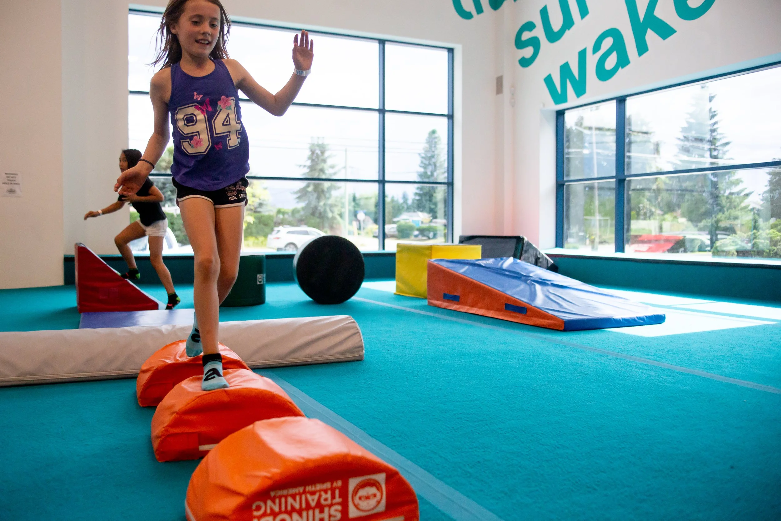 Two children playing in an indoor gym with soft foam obstacles, large windows showing trees and parked cars outside.