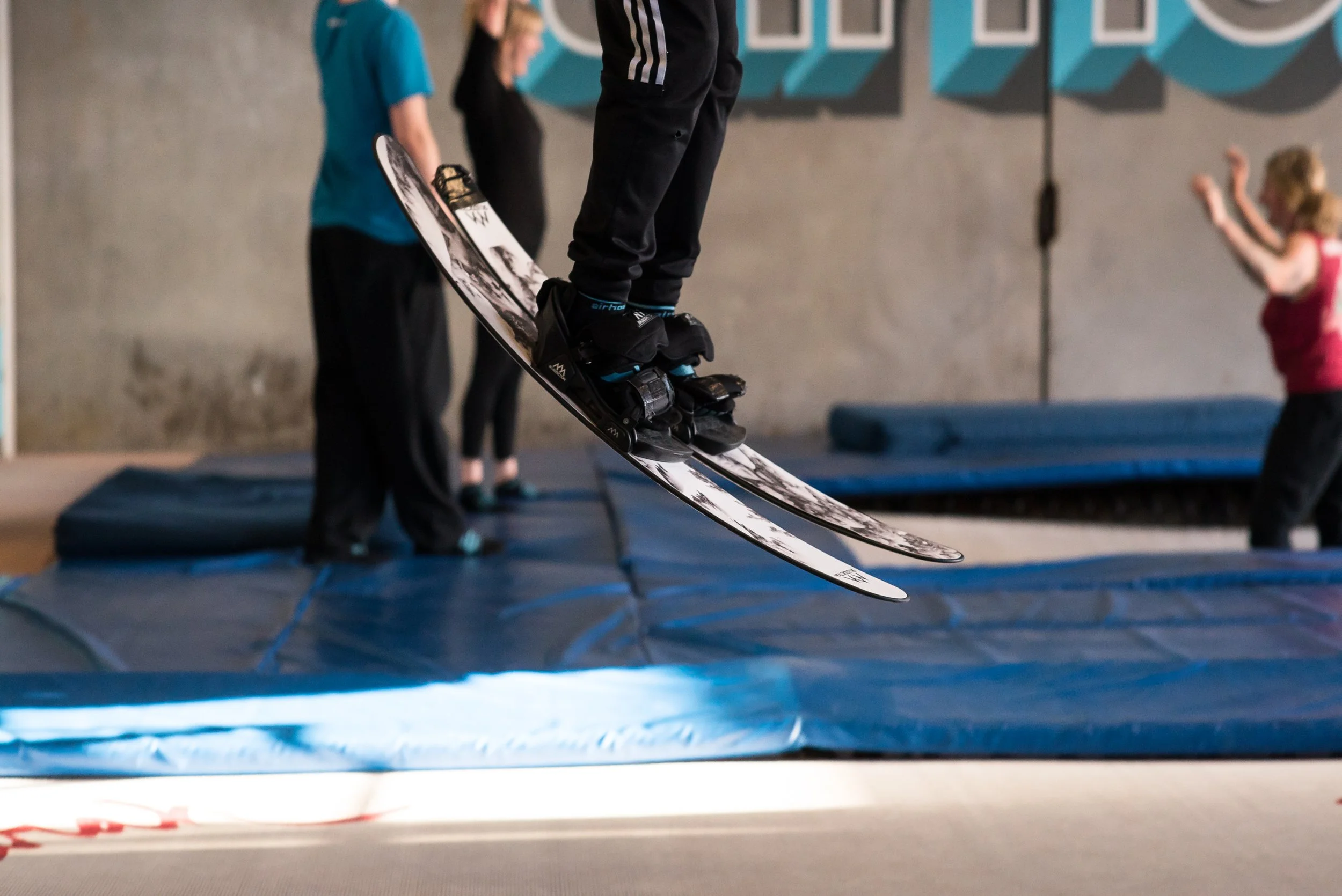 Person wearing ski gloves and pants jumping on a trampoline with skis on inside a gym, with other children in the background.