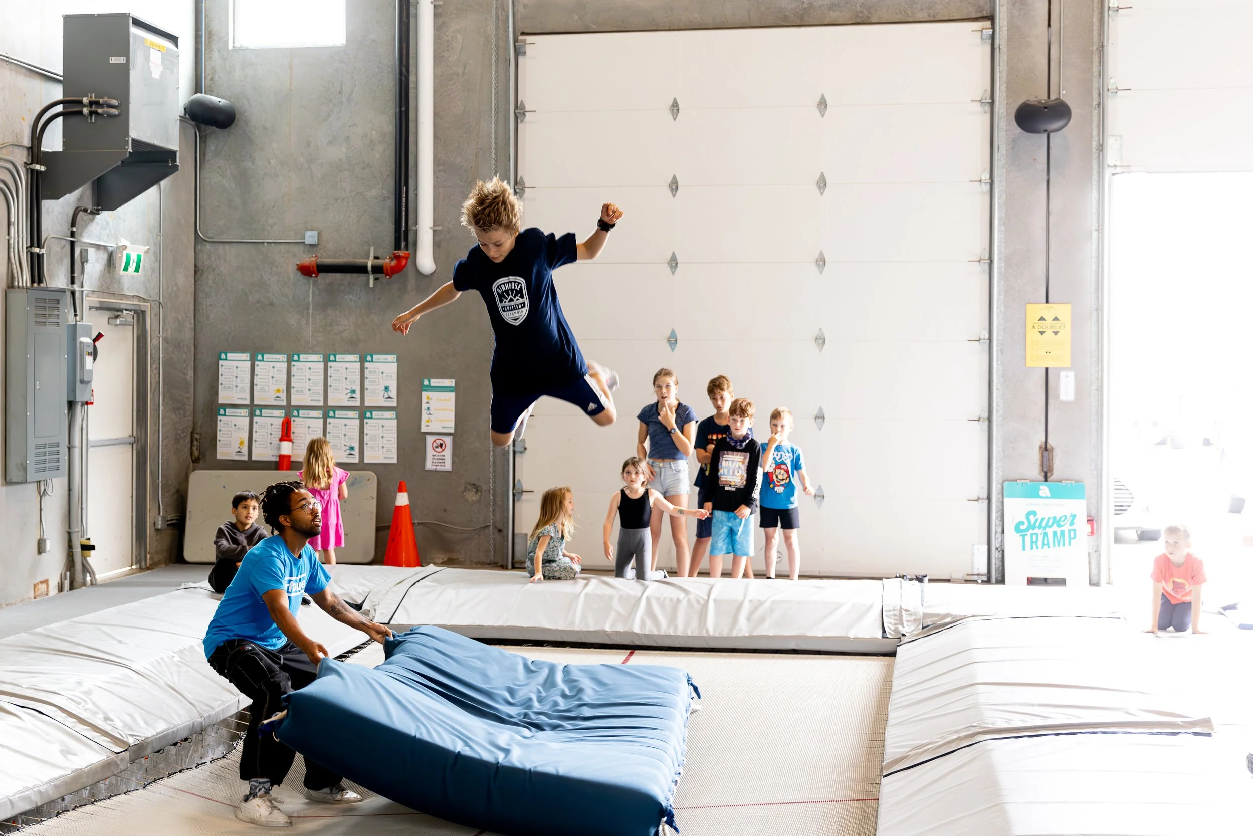 A young boy is mid-air trampoline jumping while a coach holds a large blue crash mat underneath. Several children and a woman observe in a warehouse-like indoor trampoline park with multiple mats, a white garage door, and safety signs on the wall.