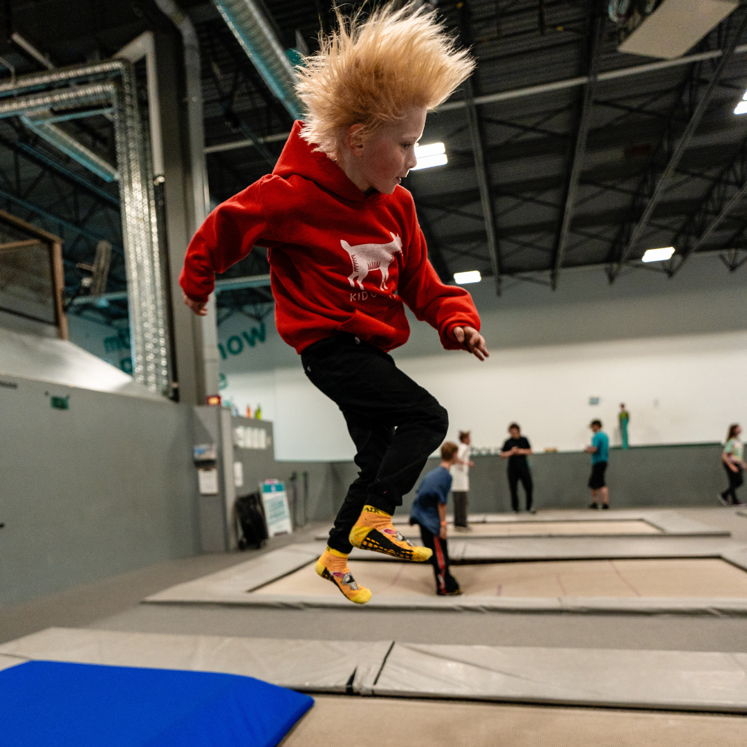 A young child with spiky blonde hair, wearing a red hoodie with a graphic of a dog and black pants, jumping on a trampoline at an indoor trampoline park. Other kids and adults are in the background.