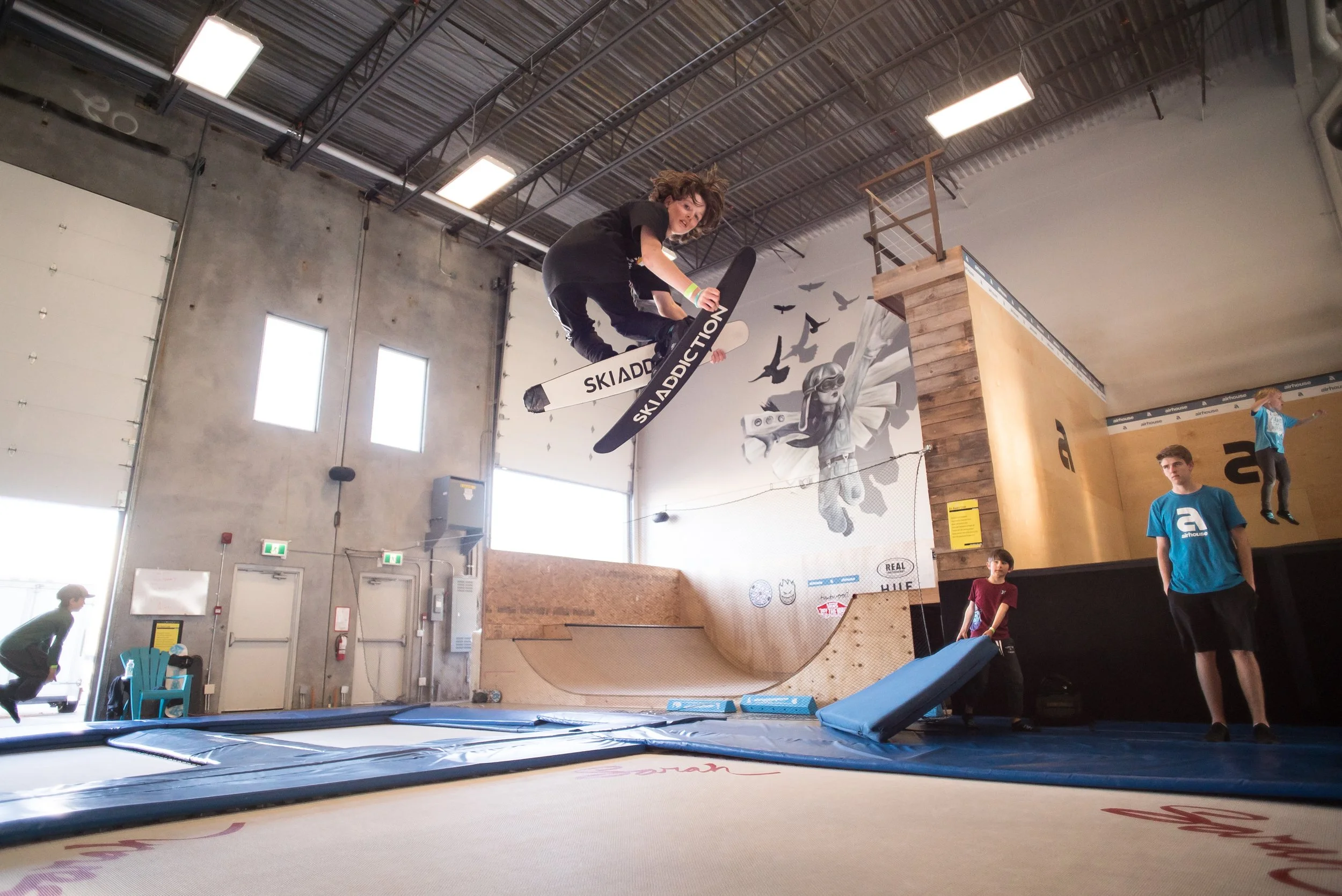 A person mid-air performing a skateboard trick at an indoor skatepark, with others watching nearby.