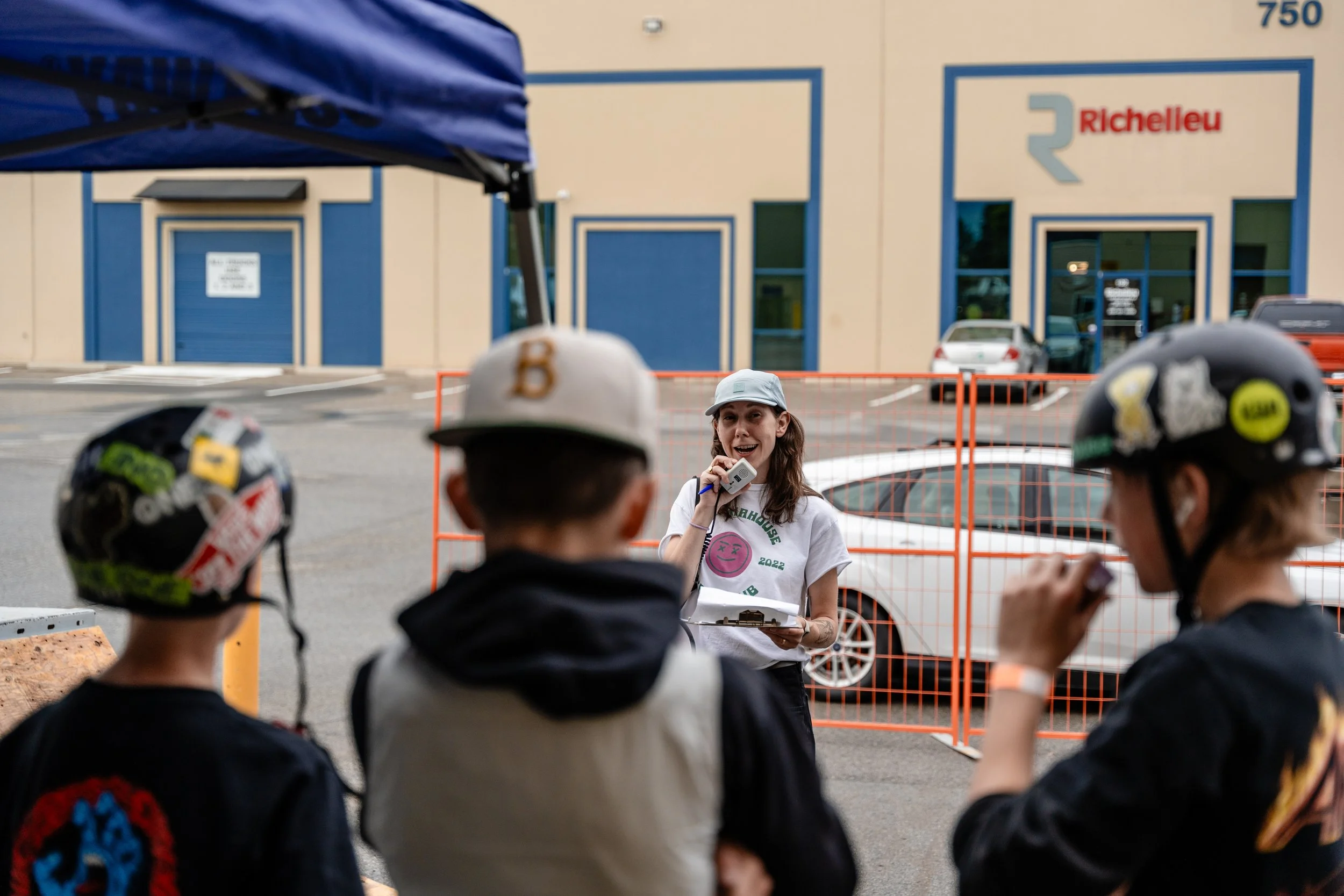 A woman speaking into a microphone while holding a clipboard, standing in front of a building with a sign that reads Richelleu, with three people with helmets in the foreground.