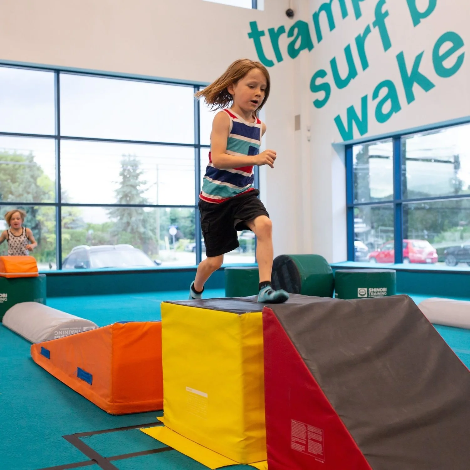 A girl jumping off a colorful foam block at an indoor play area with large windows and a sign that reads 'trampoline, surf, wake'.
