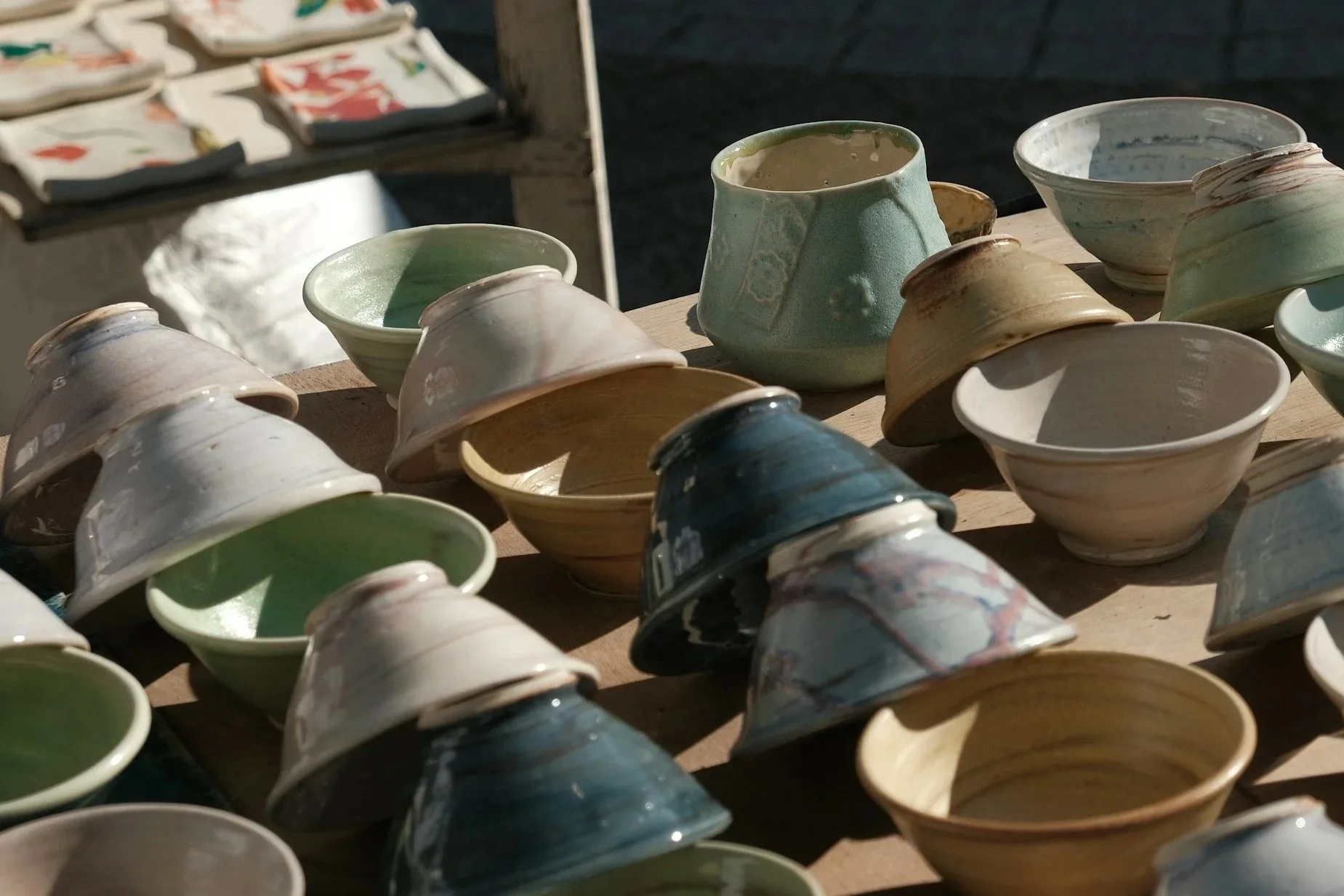 Assorted glazed ceramic bowls and cups arranged on a wooden table outdoors.