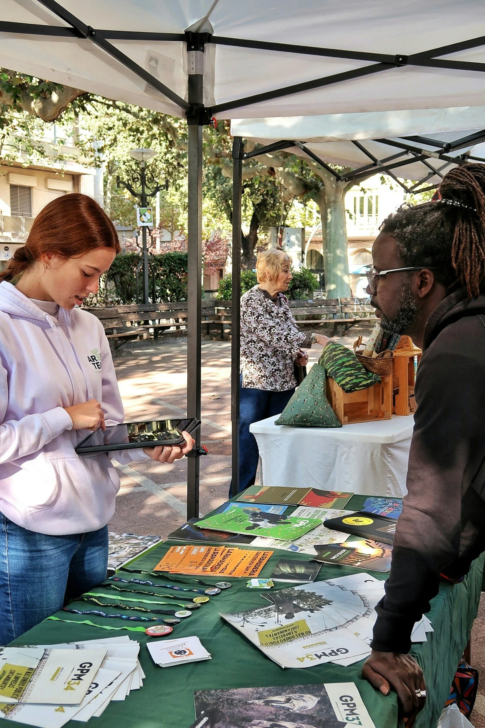 A young woman is using a tablet at an outdoor market stall with various pamphlets and buttons on the table. An older woman is behind the stall, and a man is leaning on the table, browsing the items. Trees and benches are visible in the background.